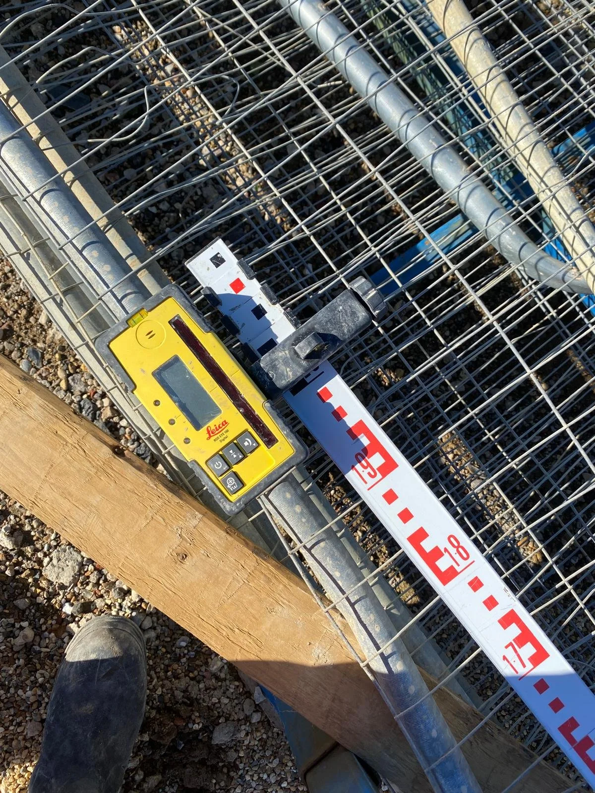 A surveyor's rotary laser level device placed on a wooden post to measure a construction fence. The device is yellow with a digital display and buttons, and is positioned near a metal fence with a measuring tape running along it.