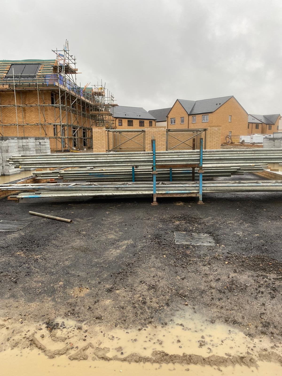 Construction site with scaffolding and building materials, residential houses in the background on a cloudy day.