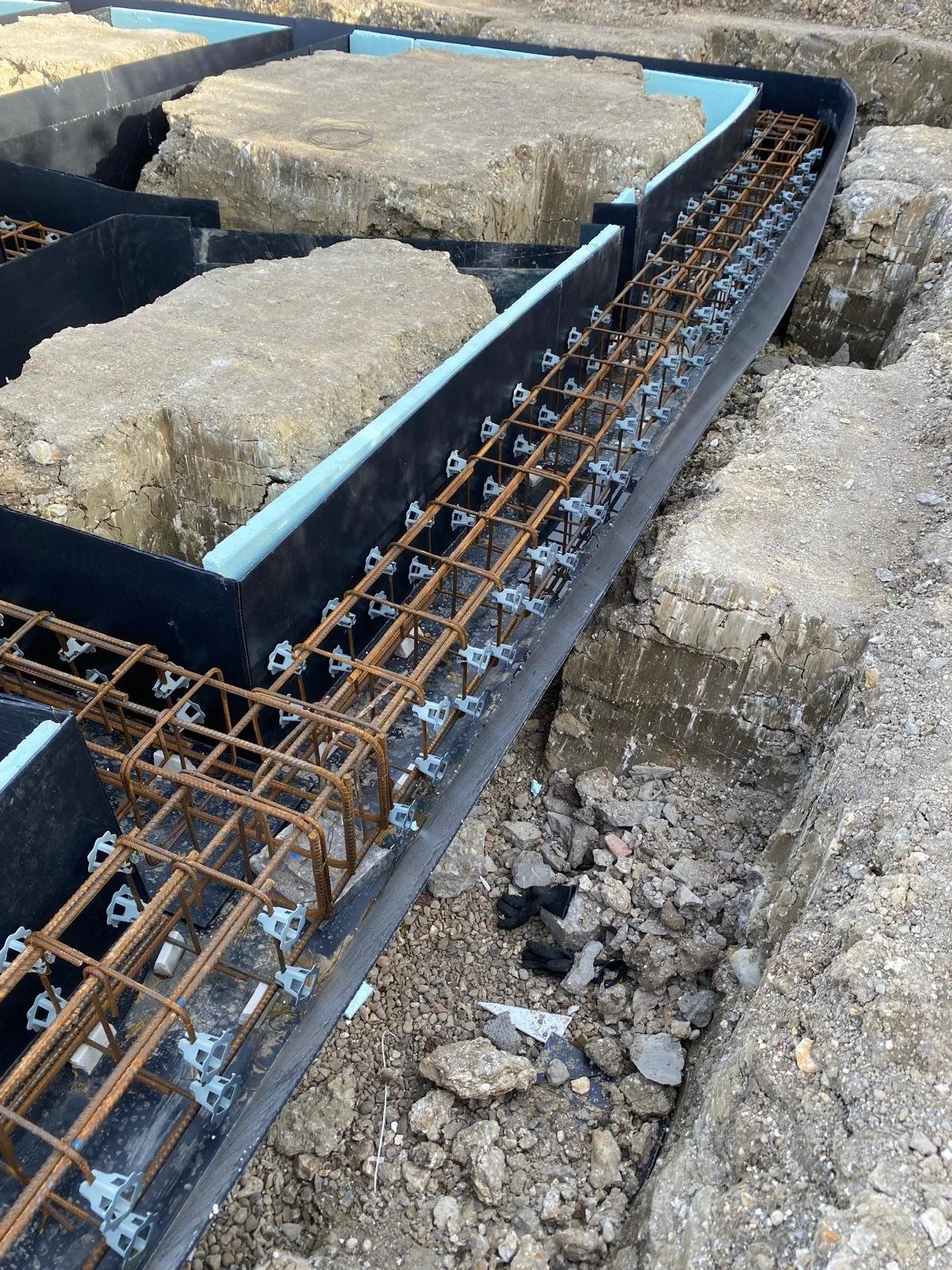 Close-up view of a construction site showing steel reinforcement bars and large rock blocks being prepared for concrete pouring. The bars are arranged within a formwork, with some of the formwork lined with black plastic sheets.