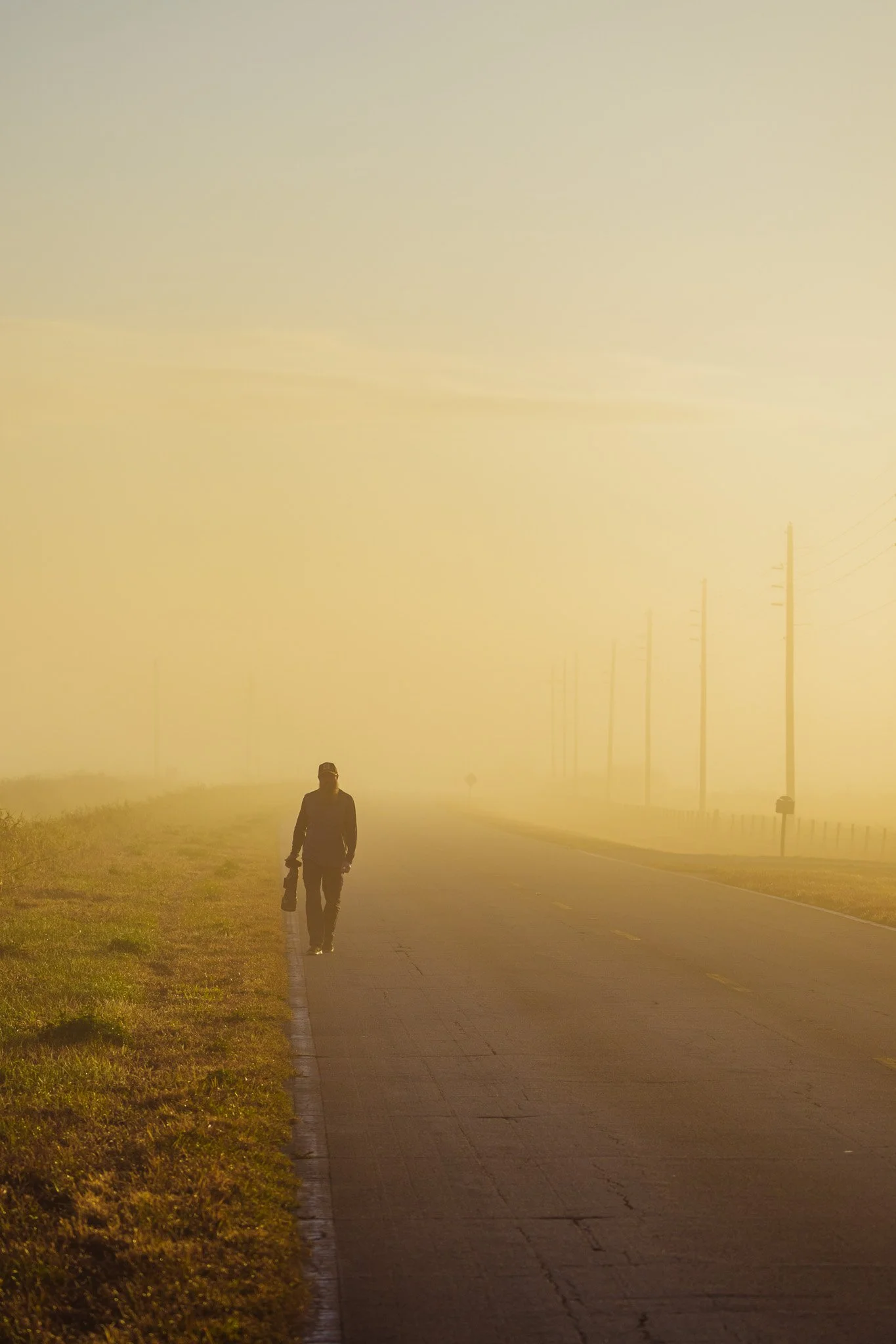 A person walking along a foggy road during sunrise or sunset, holding a bag, with power lines on the right side.