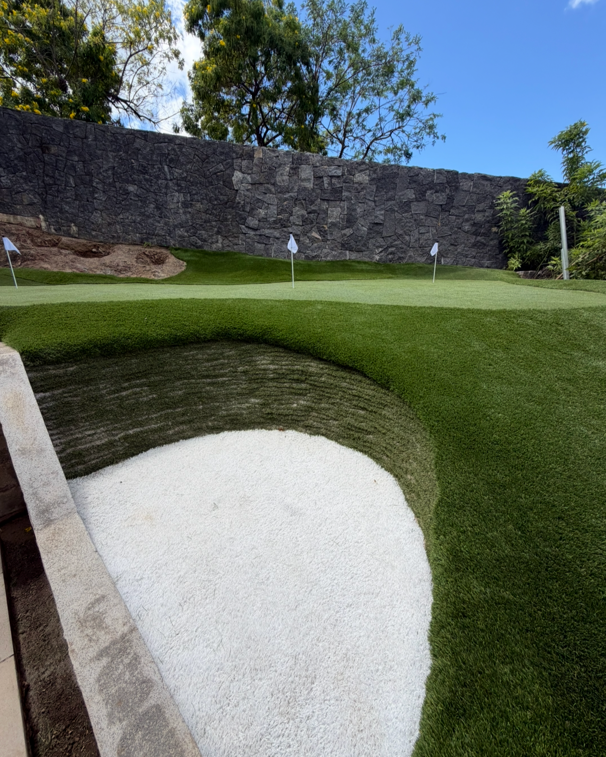 A golf course putting green with three flags, a sand bunker, and a stone wall in the background under a clear blue sky and some trees.