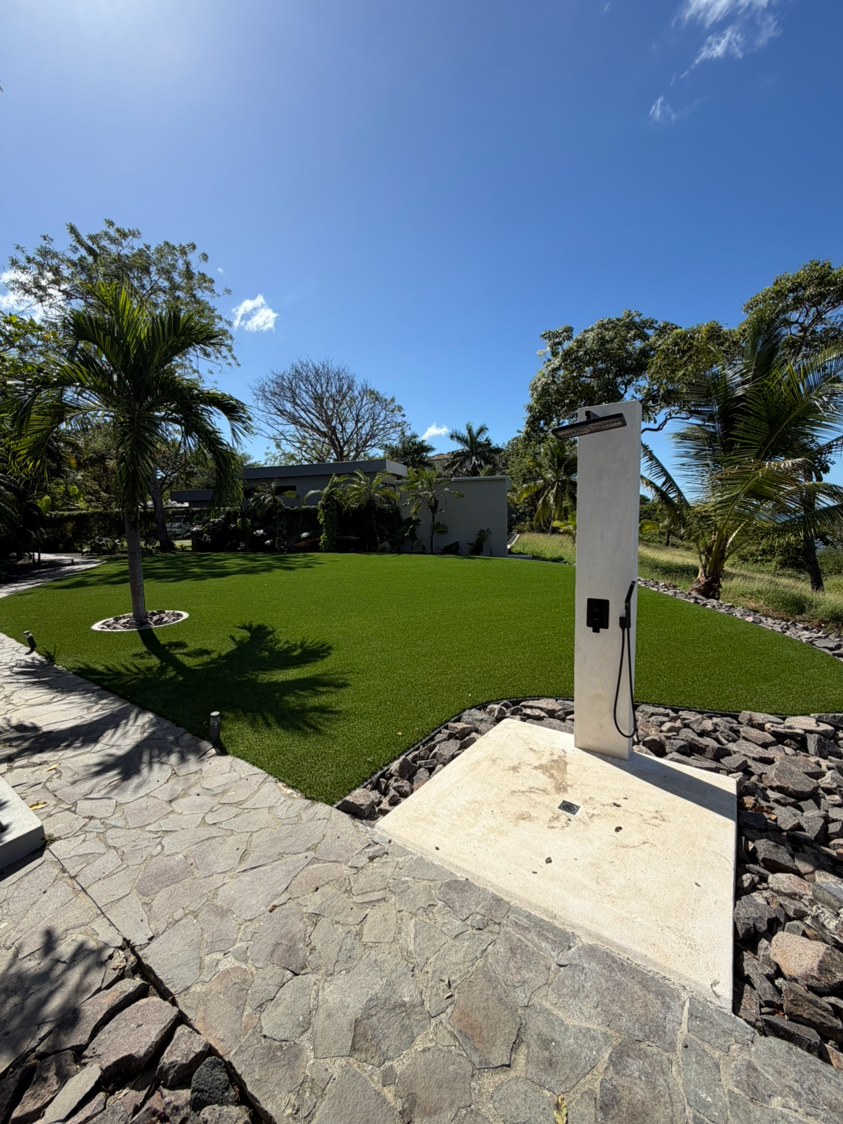 An outdoor shower in a lush garden with green grass, palm trees, and a clear blue sky.