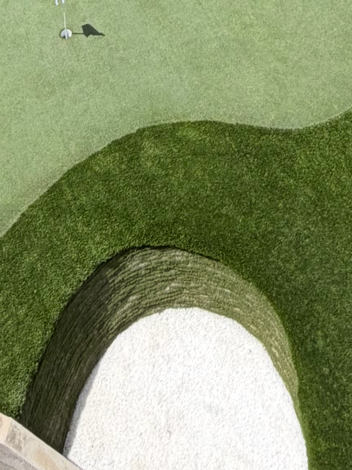 Overhead view of a golf green with a hole and flag, adjacent to a sand trap and surrounded by artificial turf.