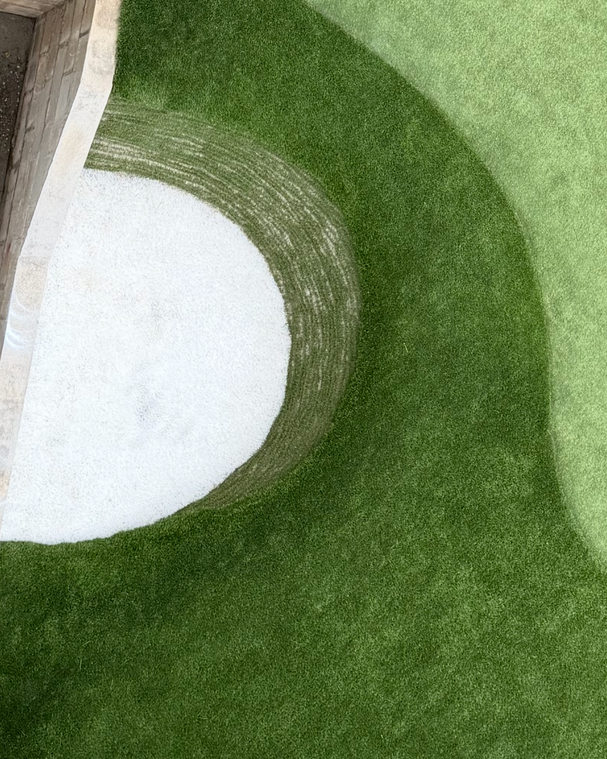 Artificial green turf with a white sand-filled circle, a golf putter, a yellow golf ball, and a white tee placed on the turf.