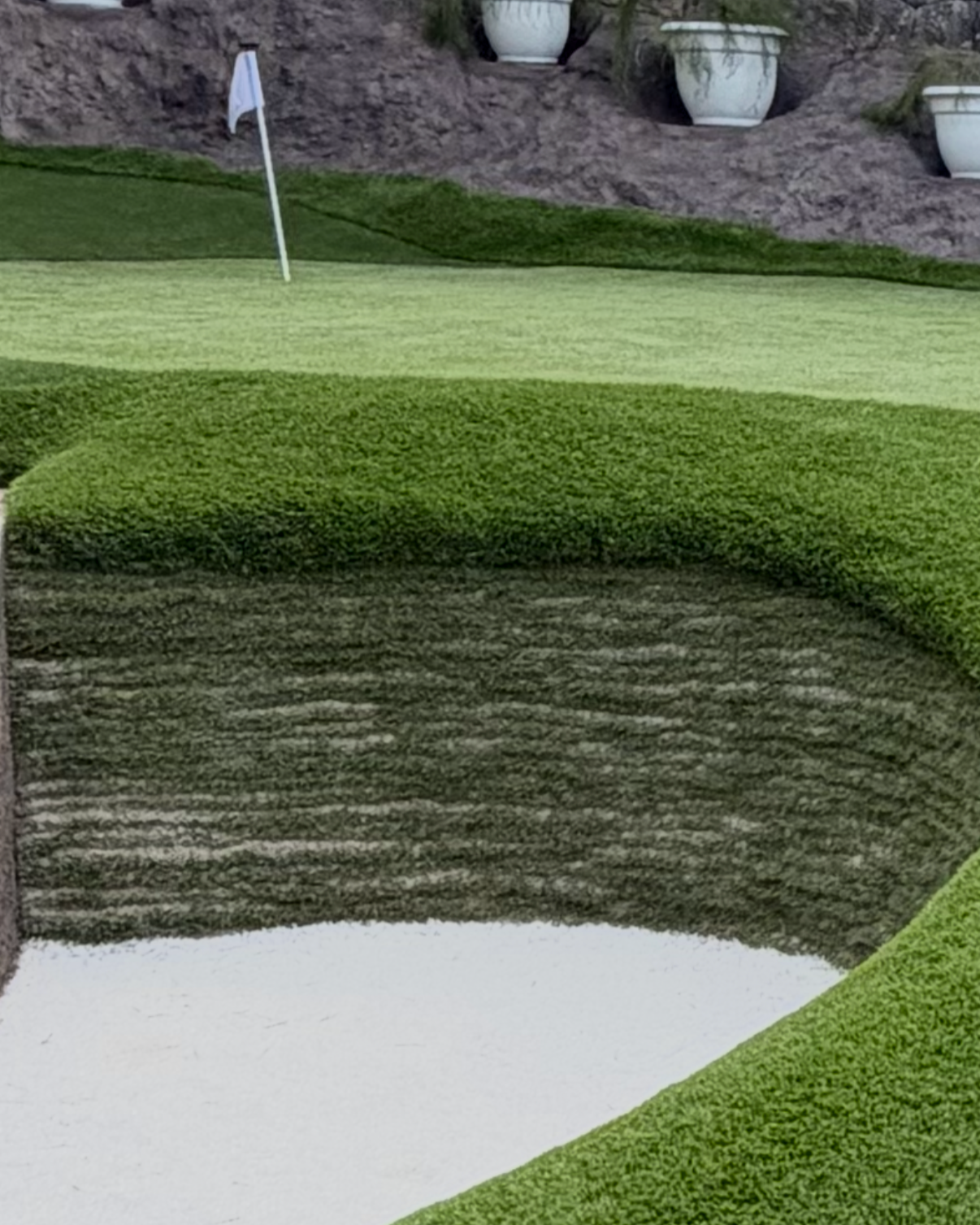 A close-up of a golf course sand trap with white sand, surrounded by well-manicured green grass and a putting green with a flag in the background, all near a rock wall with large decorative white planters.
