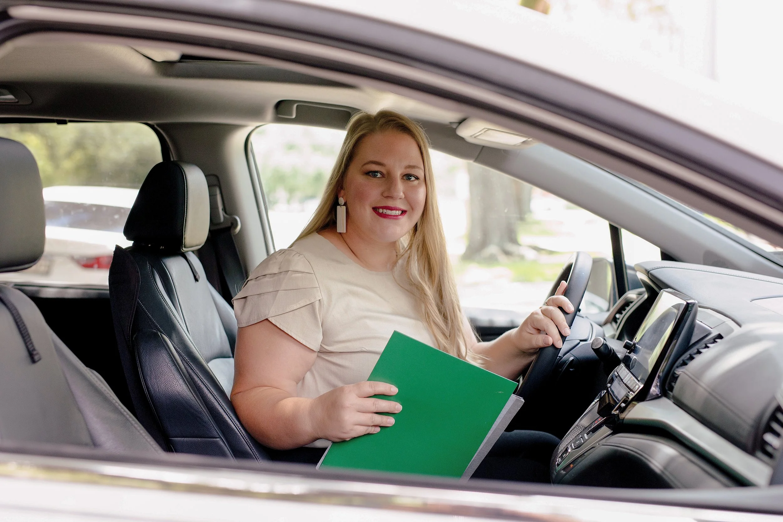 A woman sitting in the driver's seat of a car, holding a green folder and smiling at the camera.