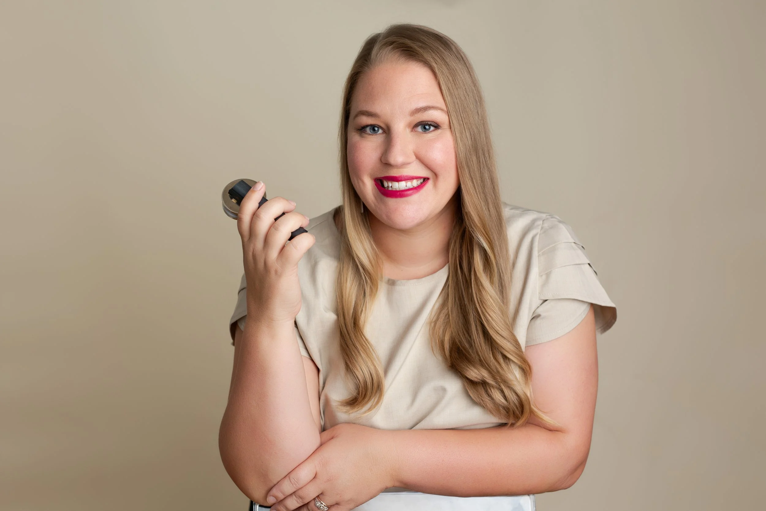 Smiling woman with long blonde hair holding a small dumbbell in front of a plain beige background.