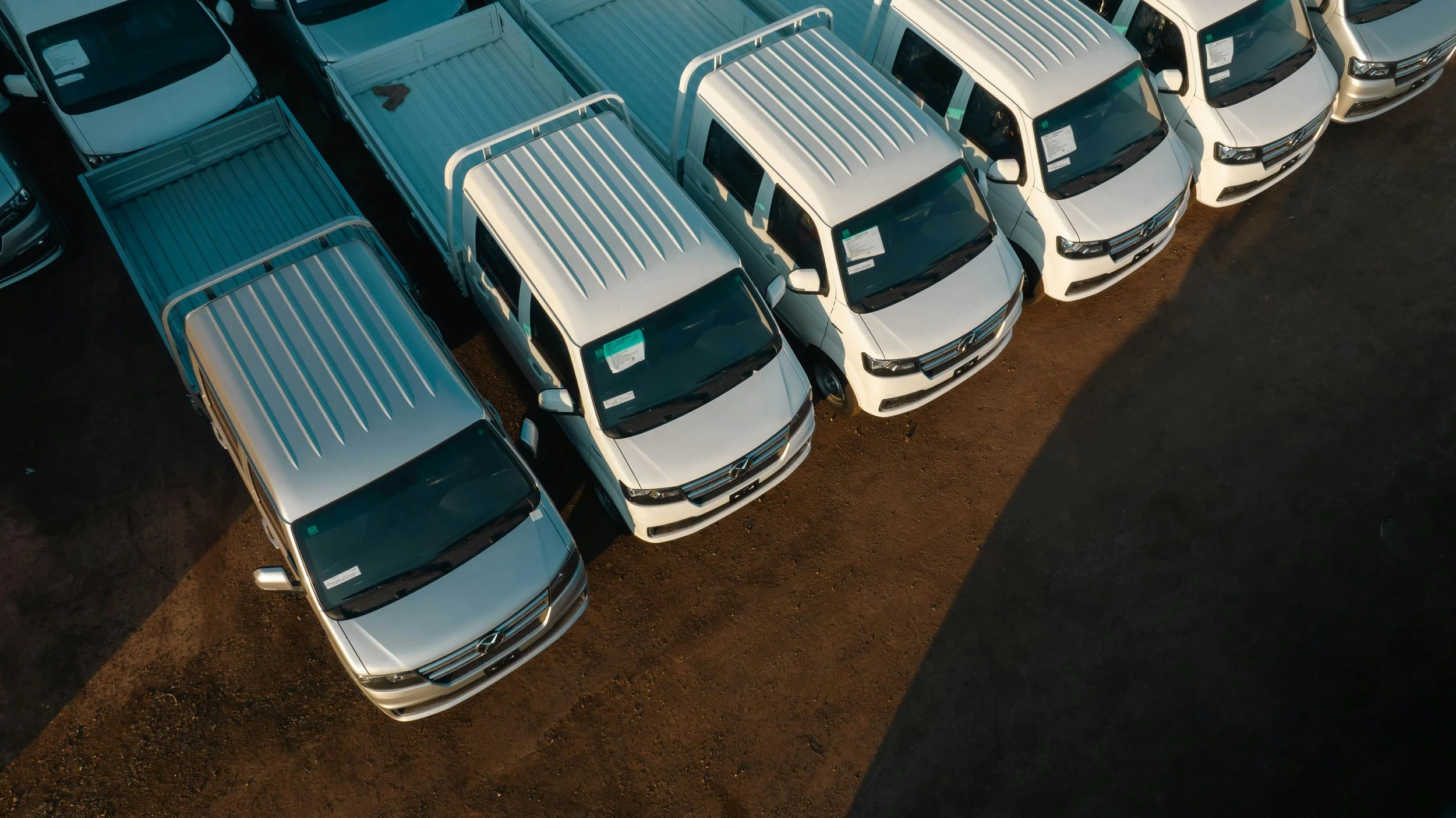 Aerial view of white pickup trucks and other vehicles parked in a lot on dirt ground, sunlight casting shadows.