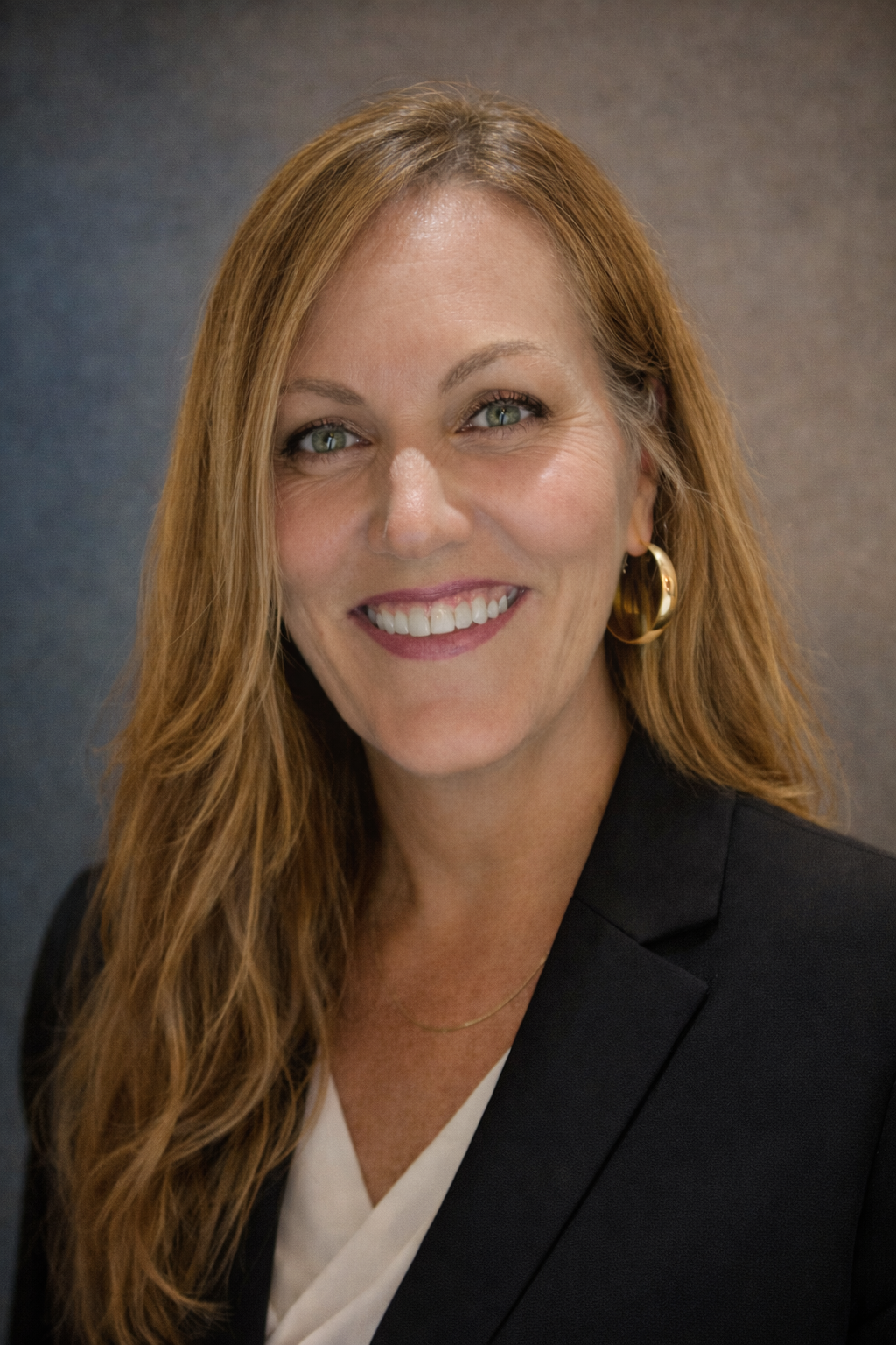 Close-up portrait of a smiling woman with long curly red hair, wearing gold hoop earrings, a black blazer, and a white top, against a dark background.