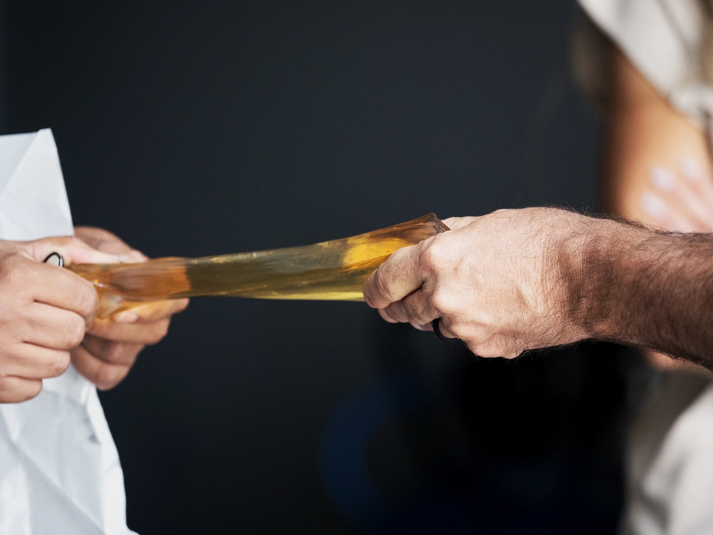 Hands exchanging a sticky, transparent substance, possibly caramel or honey, against a dark background.