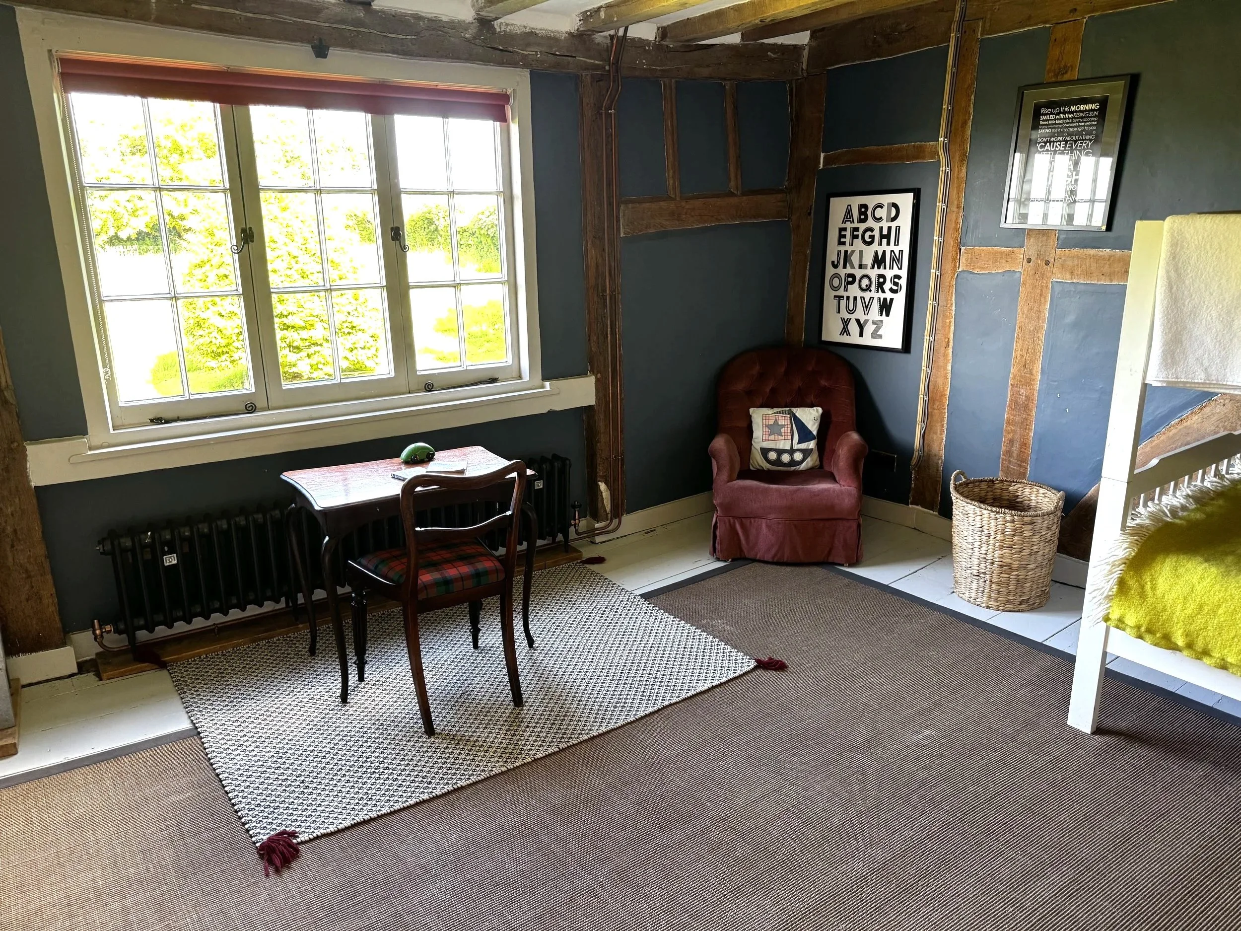 Bright room with a large window, small table with a chair, red armchair, and wooden beams on the wall, decorated with framed posters and cushions.