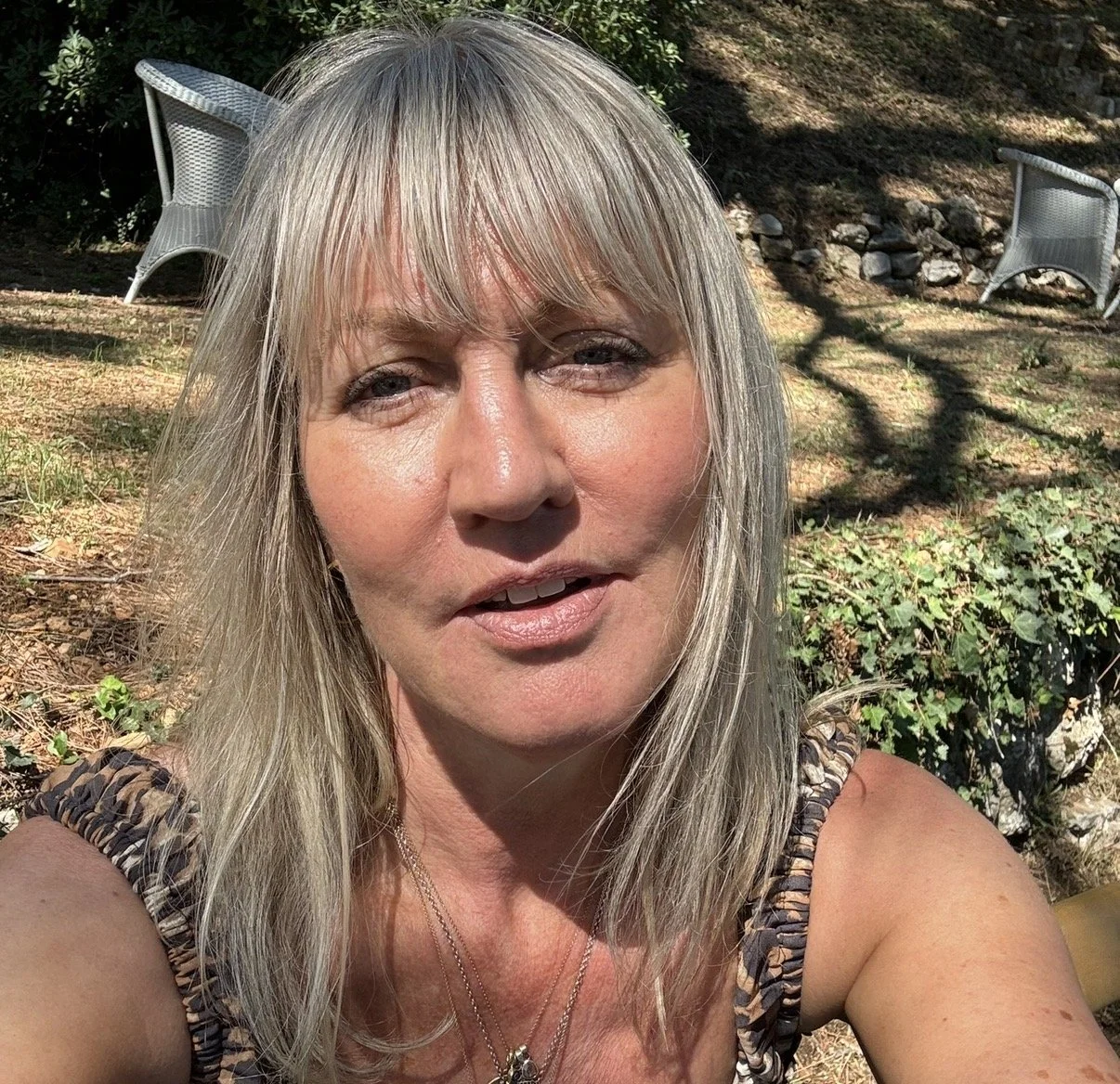A woman with long blonde hair and light makeup takes a selfie outdoors on a sunny day. She is wearing a sleeveless top with a leopard print pattern, a necklace, and has a slight smile. In the background, there are two white outdoor chairs, some greenery, and a shadow of a tree on the ground.