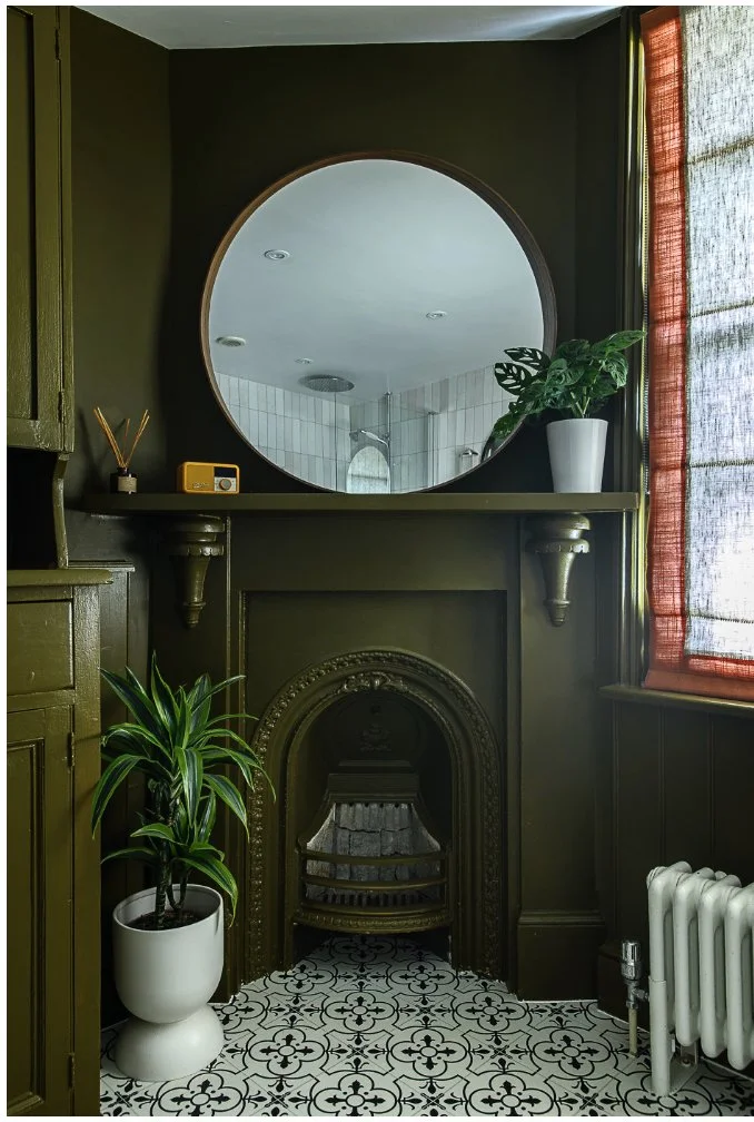 A cozy corner in a room featuring a round mirror above a vintage fireplace mantle, a potted plant on the mantle, a larger potted plant on the floor, patterned black and white tile flooring, a window with red-orange curtains, and a white radiator.
