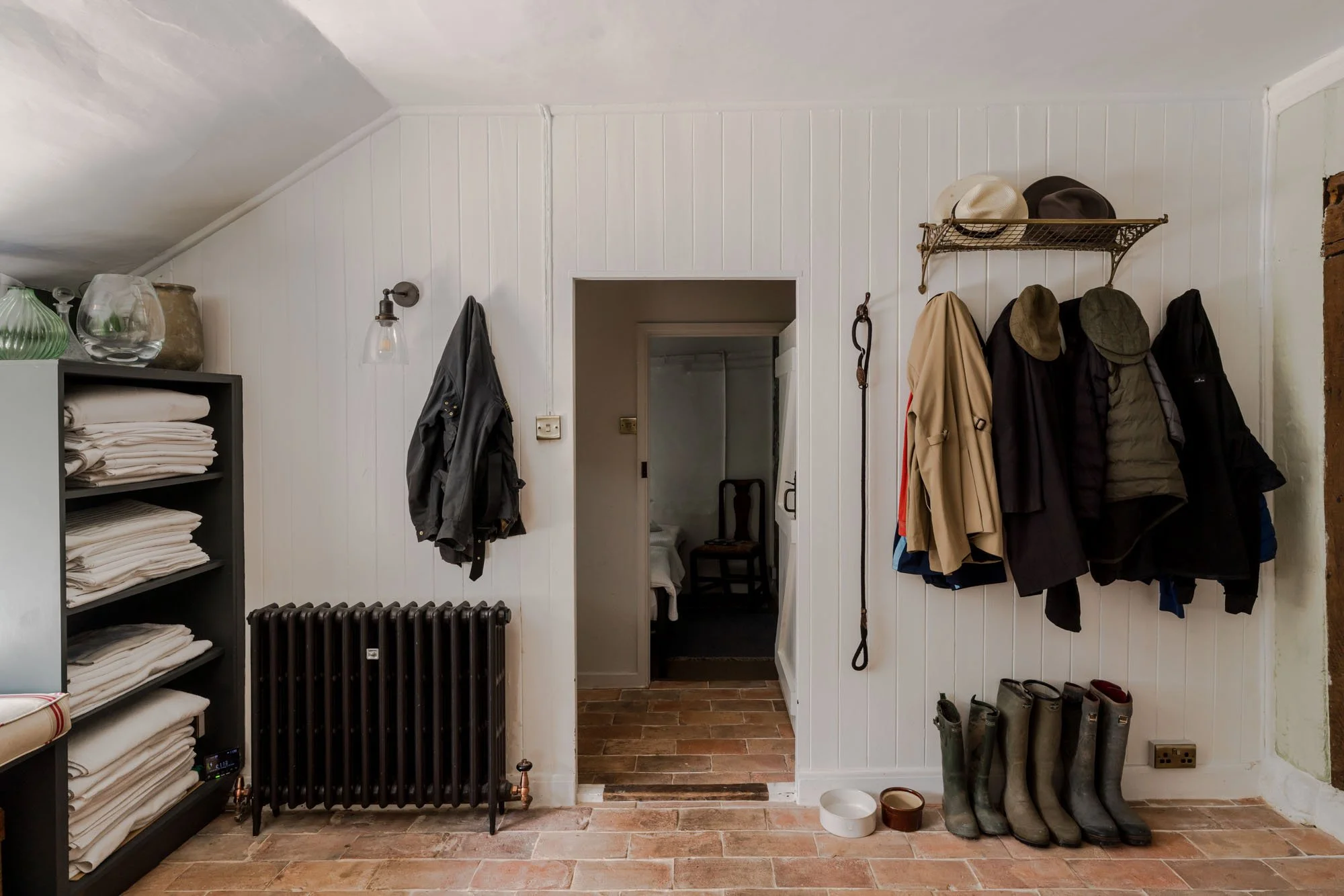 Entryway with coat rack holding jackets, hats, and umbrellas, black radiator, shelving with folded linens, and boots on the floor.