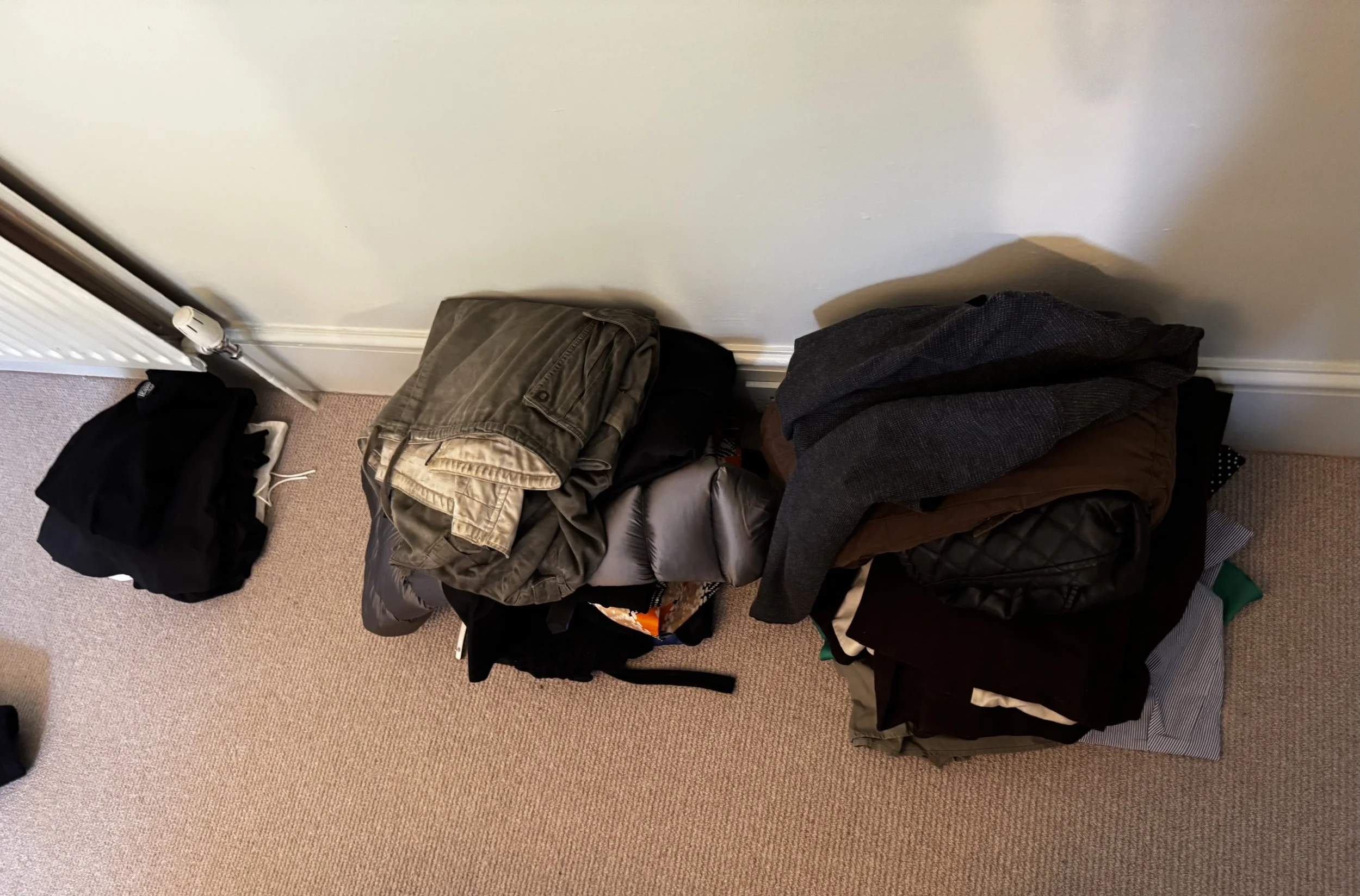 Pile of clothes and bags on a beige carpeted floor next to a white wall and radiator.