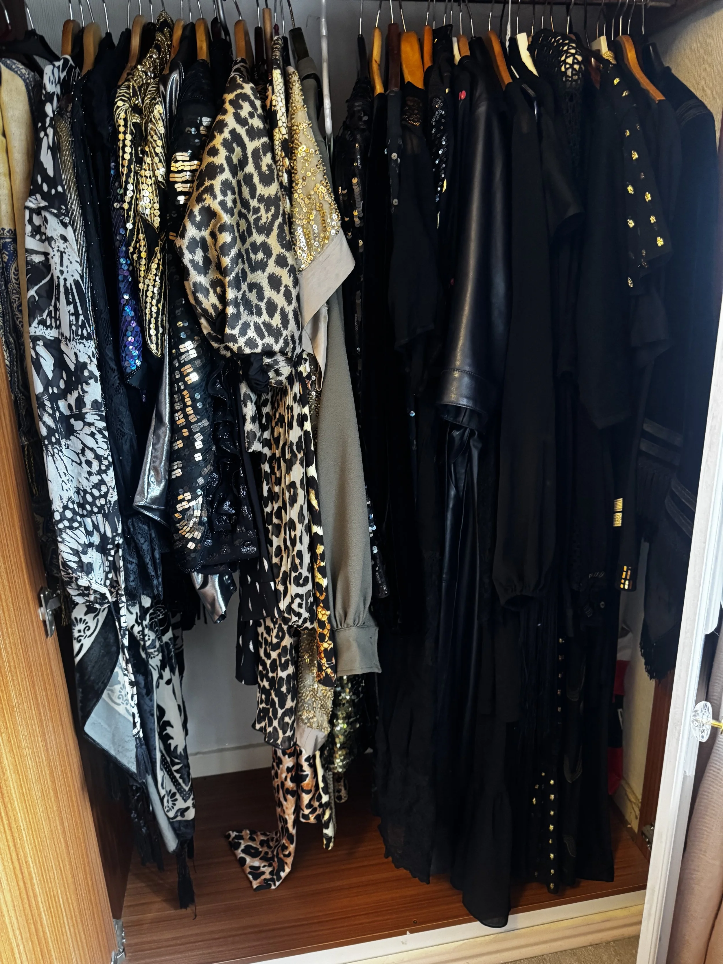 A closet with a collection of black, gold, and animal print clothing items hanging on wooden hangers, including dresses, jackets, and tops.