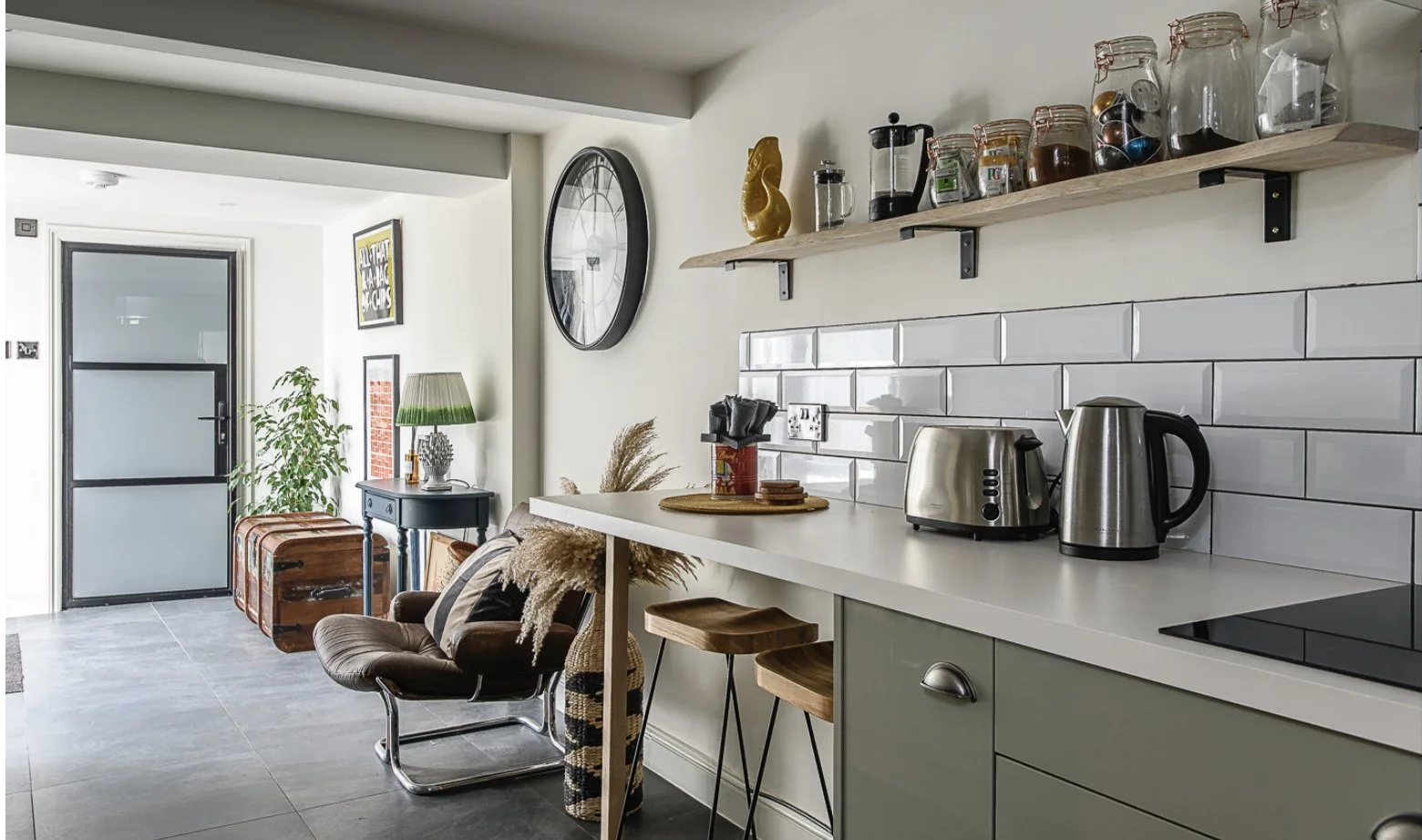 Interior view of a modern kitchen and living area with white subway tile backsplash, stainless steel appliances, and open shelving with jars and spices; living space features a brown chair, side table with a lamp, and decorative elements.