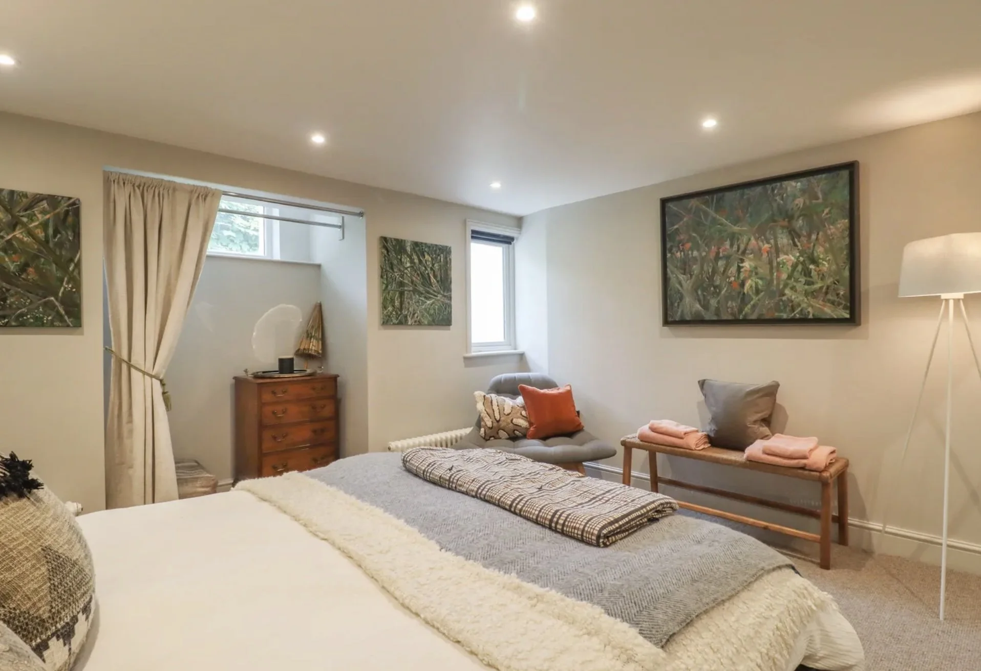 Cozy bedroom with a bed, bench, and two chairs, decorated with artwork and pillows, and illuminated by indoor lighting.