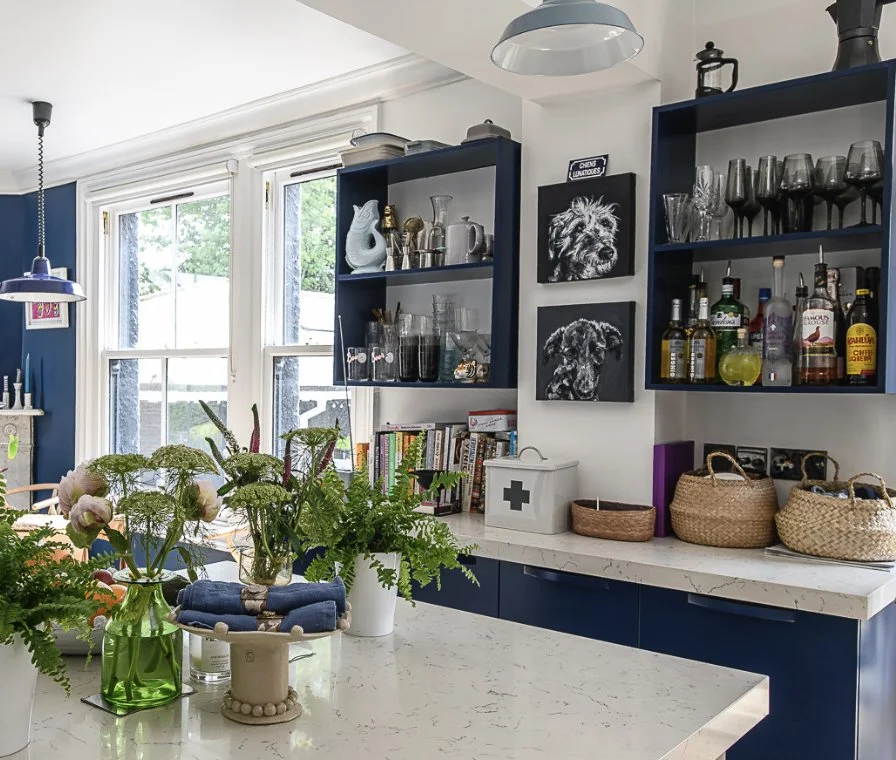 Bright kitchen with white marble countertop, open blue shelves holding glassware, liquor bottles, and decorative items, and a large window with greenery outside. There are fresh flowers and plants on the counter.