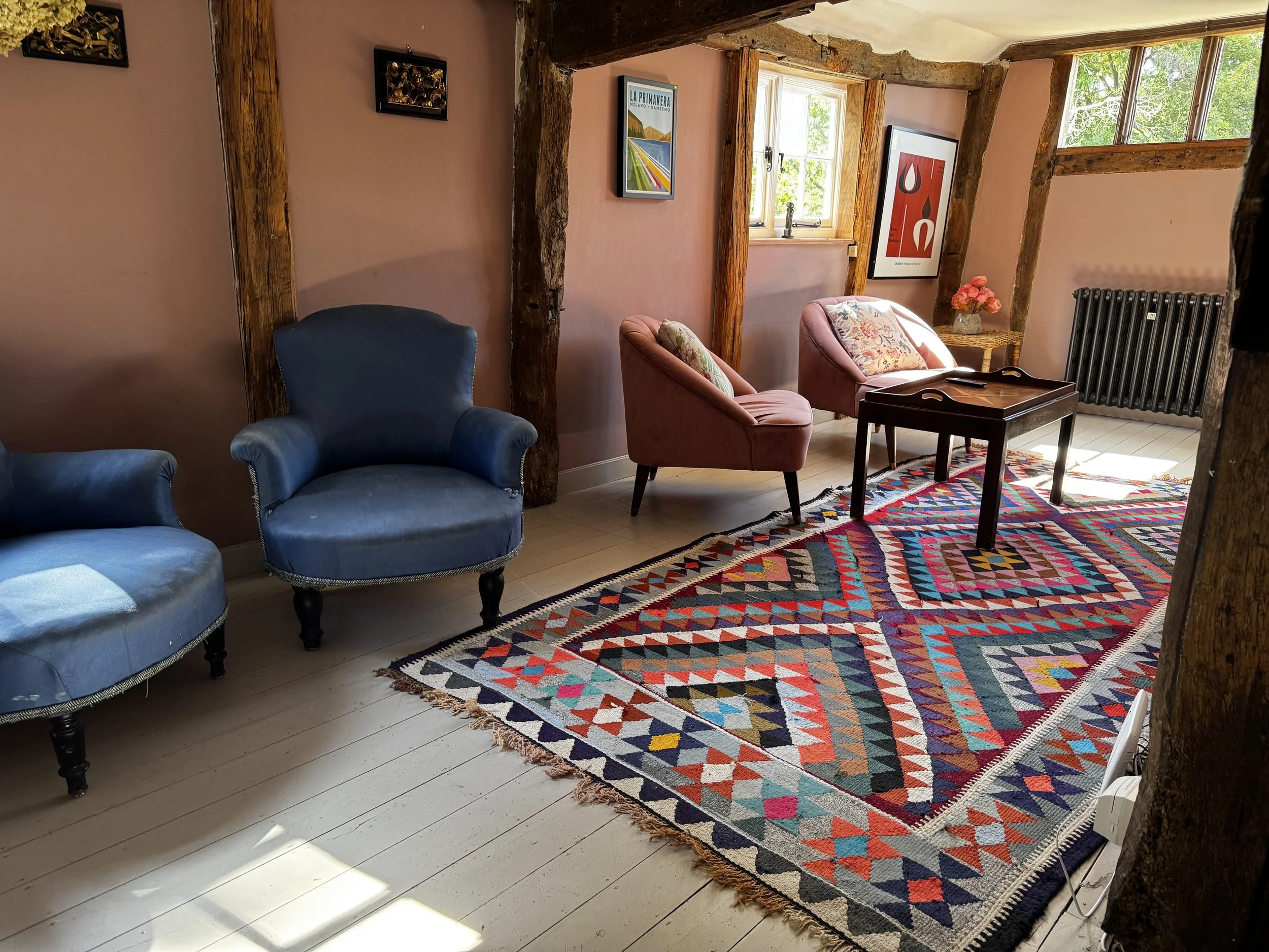A cozy living room with pink walls and exposed wooden beams, featuring two vintage blue armchairs, two pink velvet chairs with floral cushions, a colorful geometric patterned rug, a small wooden side table with pink flowers, framed artwork on the wal
