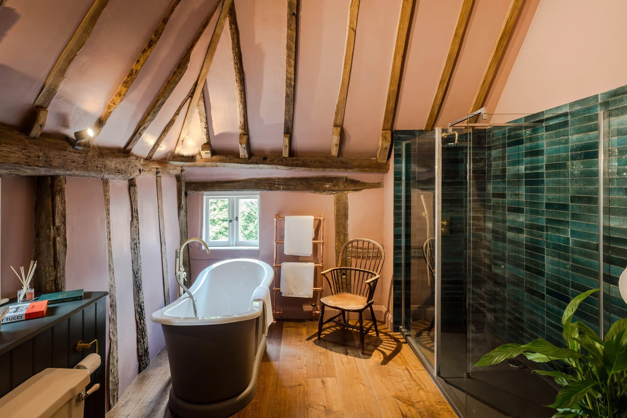 Bathroom with a freestanding bathtub, wooden beams on ceiling, small window, glass shower enclosure with green tiles, wooden chair, towel rack, and a small wooden table with a plant.