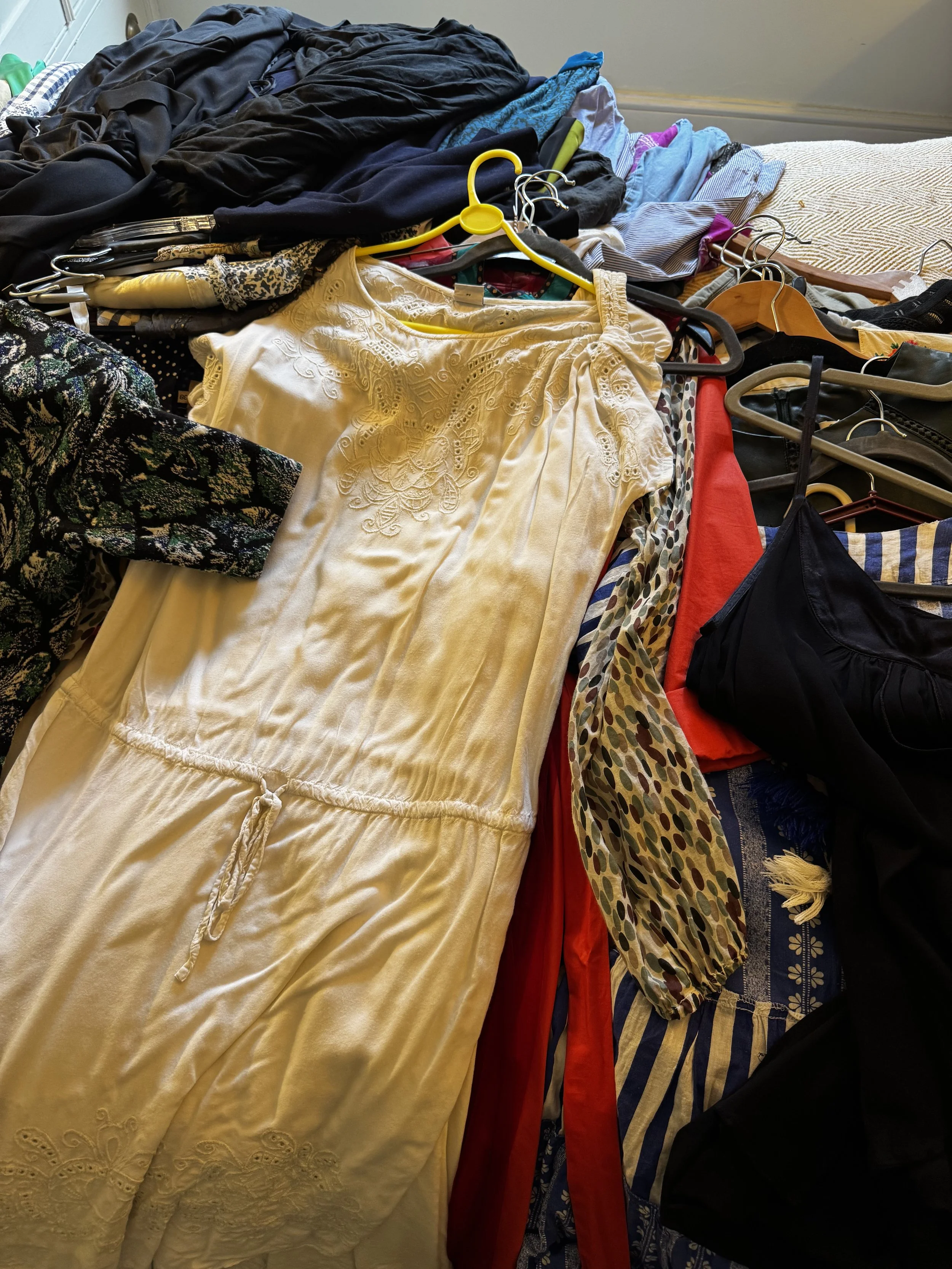 A pile of various clothes, including a yellow dress, black, blue, and patterned garments, with some hangers on top, laid out on a bed.