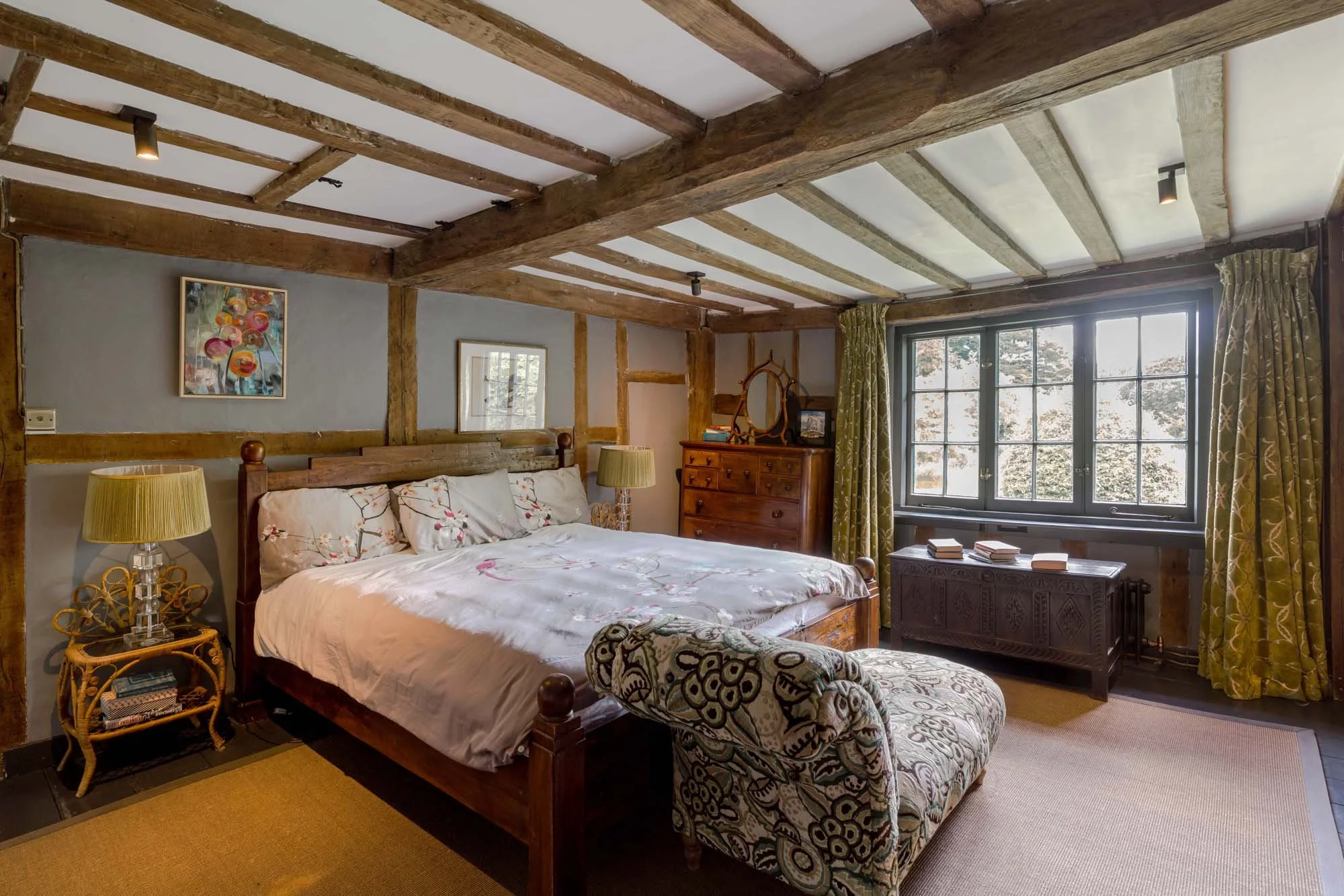 A cozy bedroom with wooden beam ceilings, featuring a large bed with floral bedding, a patterned bench at the foot of the bed, and a large window with green curtains showing trees outside. There are two bedside lamps, a wooden dresser with a mirror, 