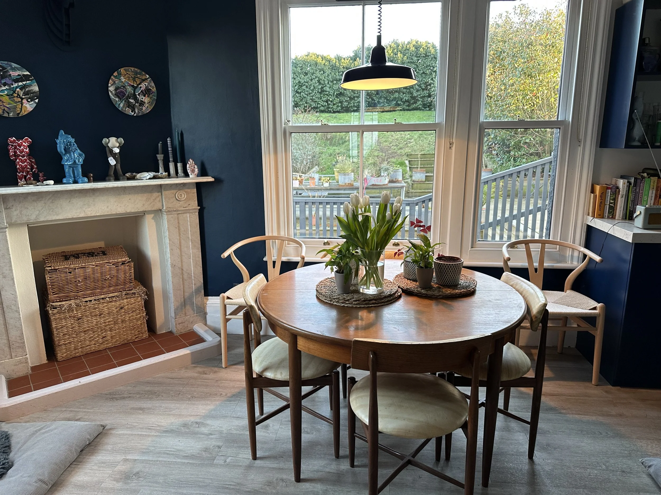 A cozy dining room with a round wooden table set near large windows, decorated with a vase of white tulips and smaller potted plants, surrounded by white chairs, with a blue accent wall and a fireplace decorated with art and figurines.