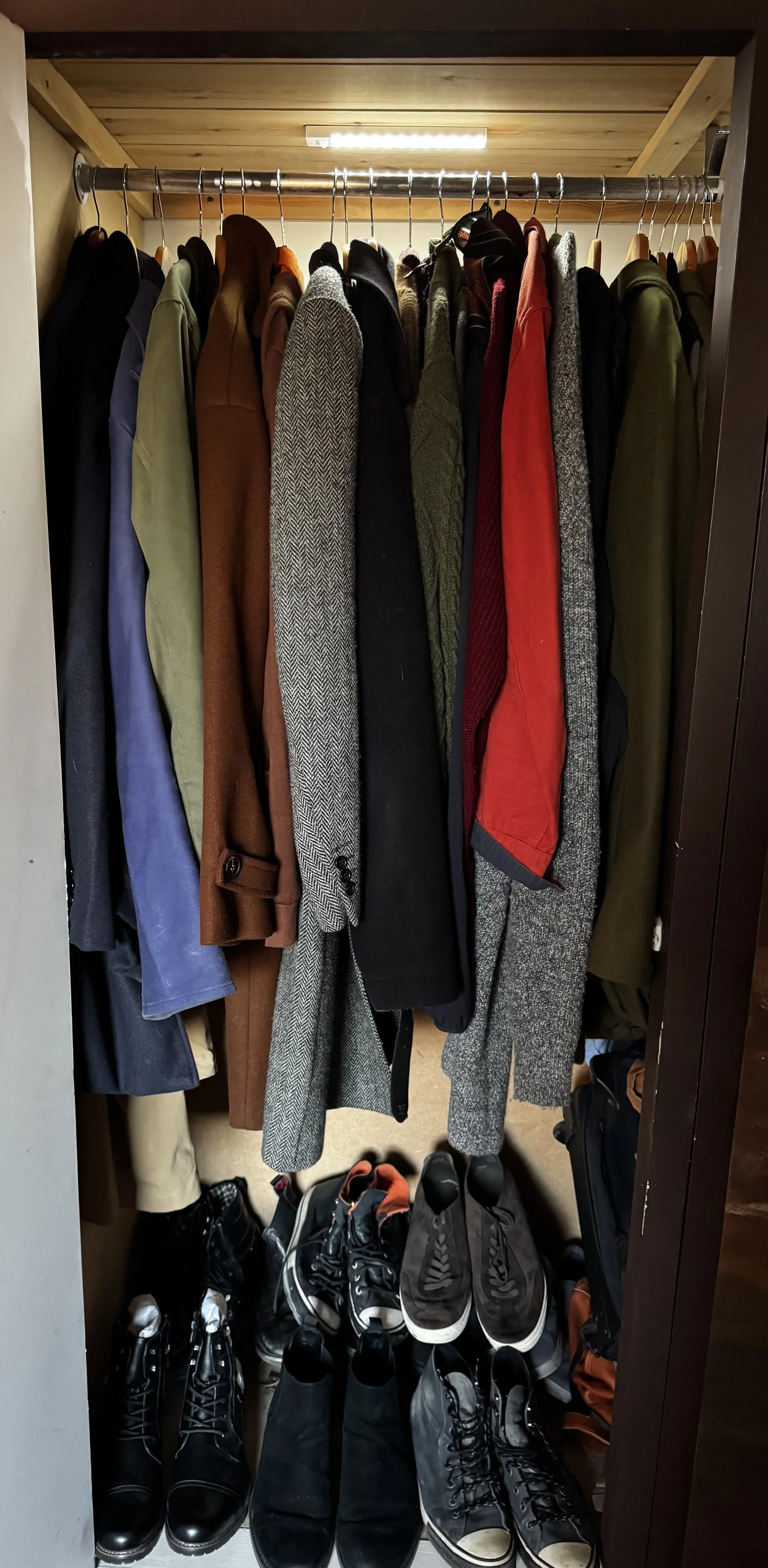 A closet with a hanging rod filled with various jackets, coats, and blazers in different colors and styles, and shoes on the floor below, including boots, sneakers, and dress shoes.