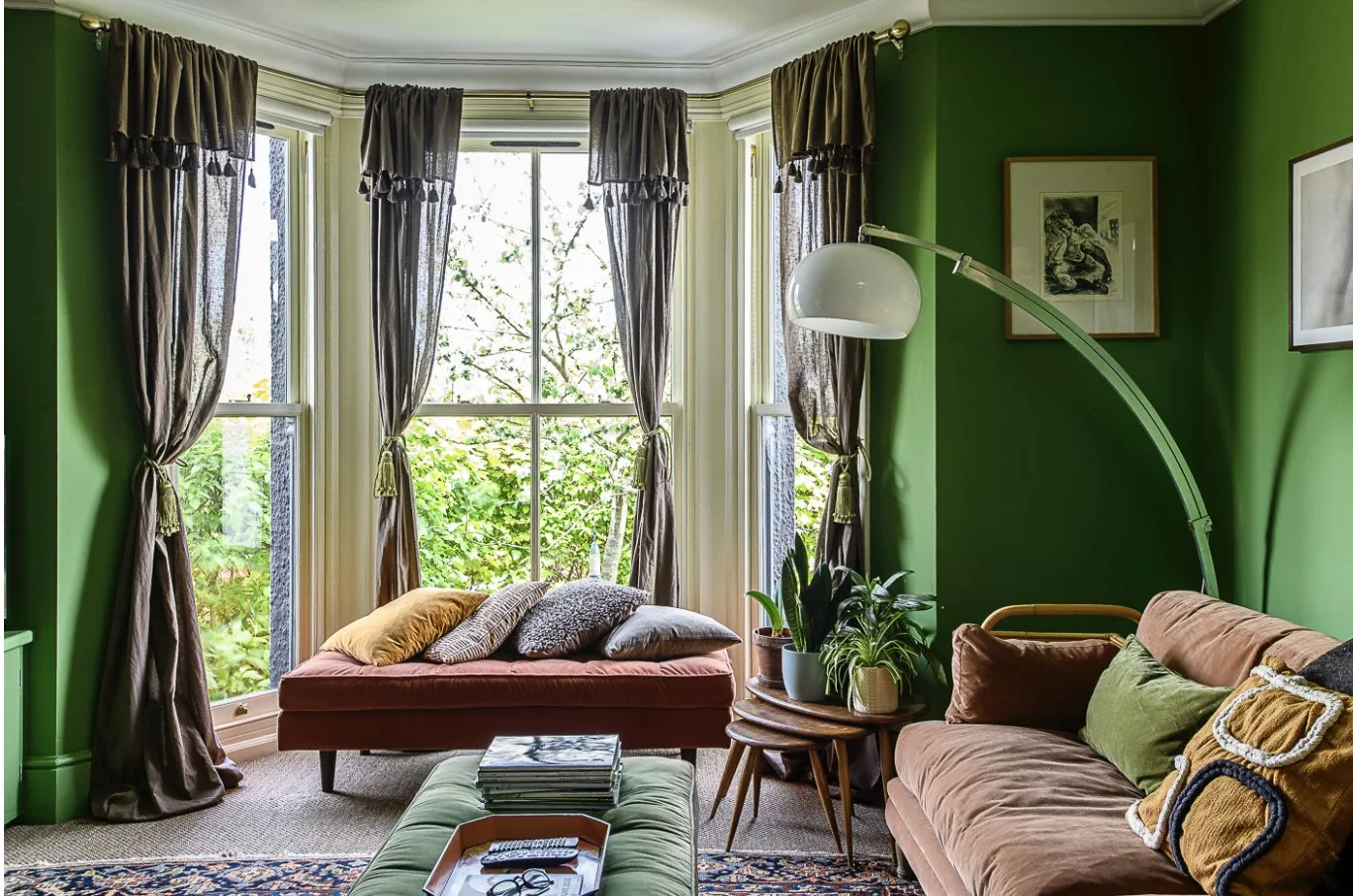 Living room with green walls, large bay window with gray curtains, brown sofa with cushions, side table with potted plants, framed artwork on wall, floor lamp with white shade, coffee table with magazines, and a patterned rug.