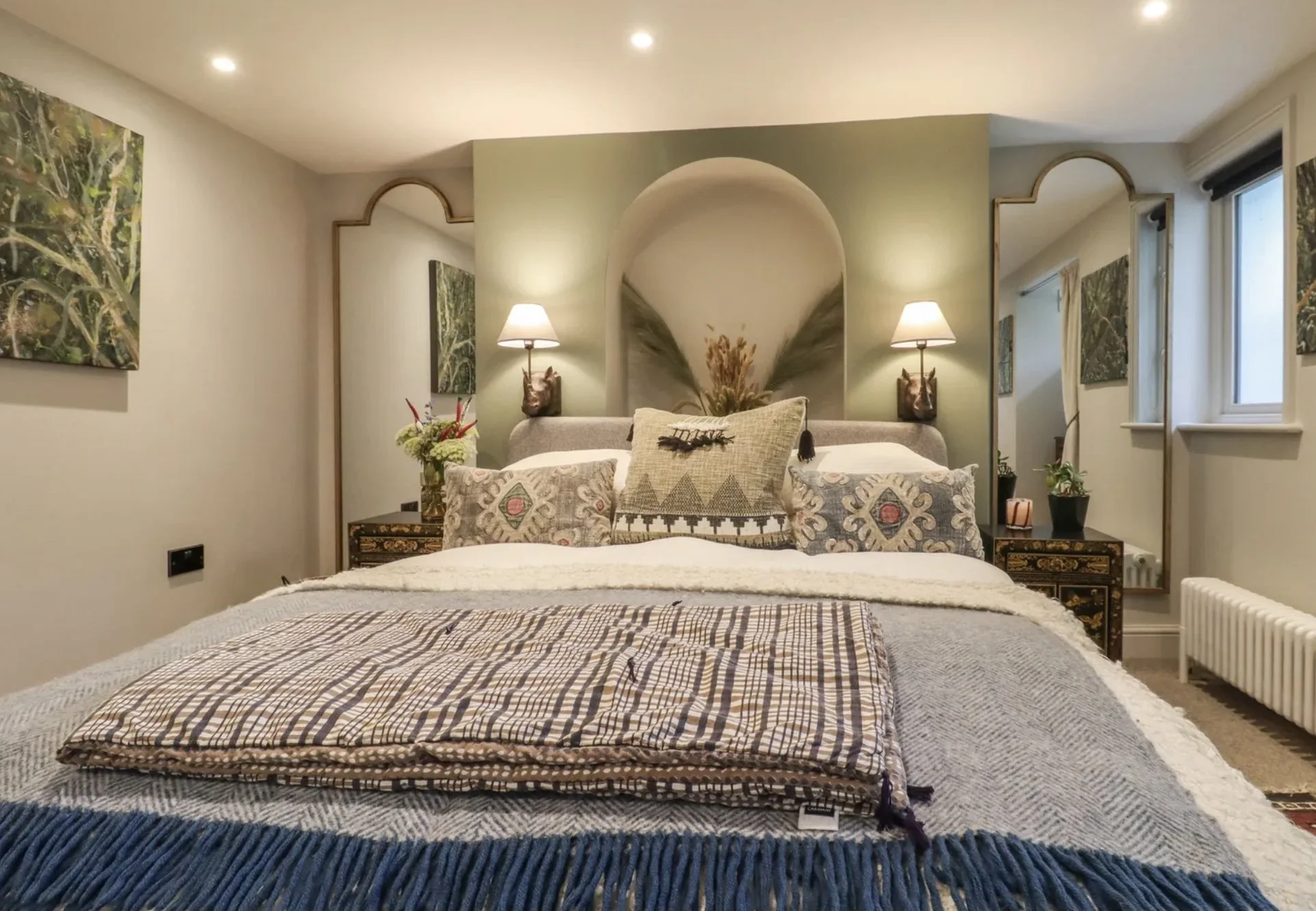 A cozy bedroom with a bed featuring patterned pillows, a textured blanket, and a decorative throw. There are two nightstands with lamps, plants, and a flower arrangement. Behind the bed, a green accent wall has a framed reflection and is flanked by m