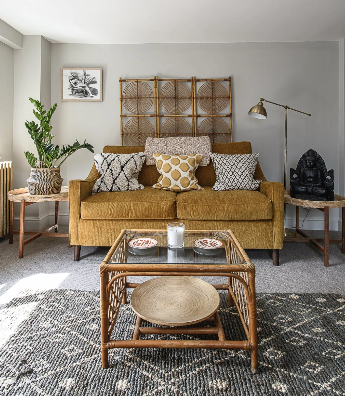 Living room with a mustard-colored sofa adorned with patterned cushions, a wooden coffee table with decorative plates and a candle, a side table with a potted plant, a black sculpture, a standing brass lamp, and a wall art piece, all on a textured ru