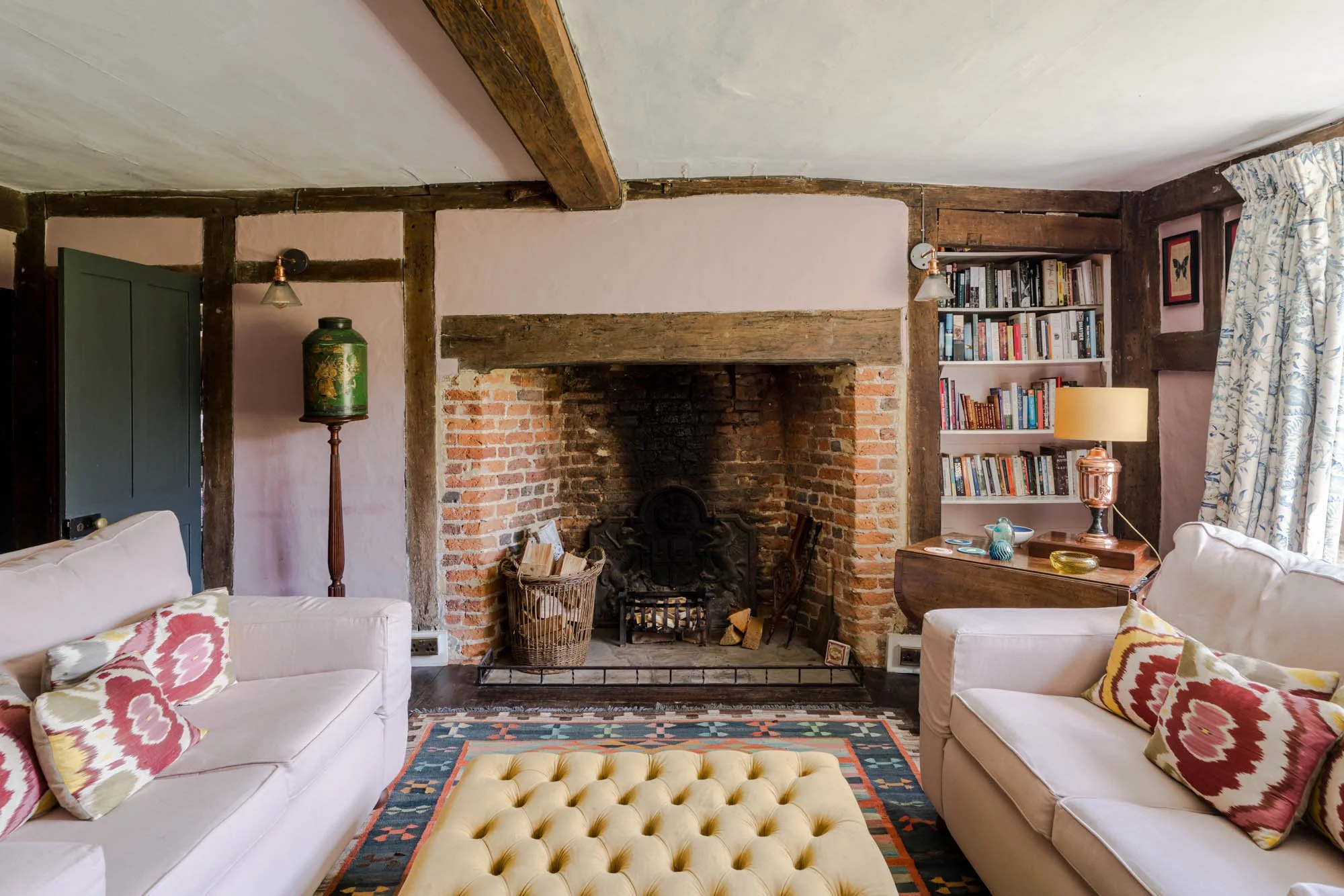 A cozy living room with a brick fireplace, two white sofas with colorful pillows, a yellow tufted ottoman, a bookshelf filled with books, a side table with a lamp, and a patterned rug.