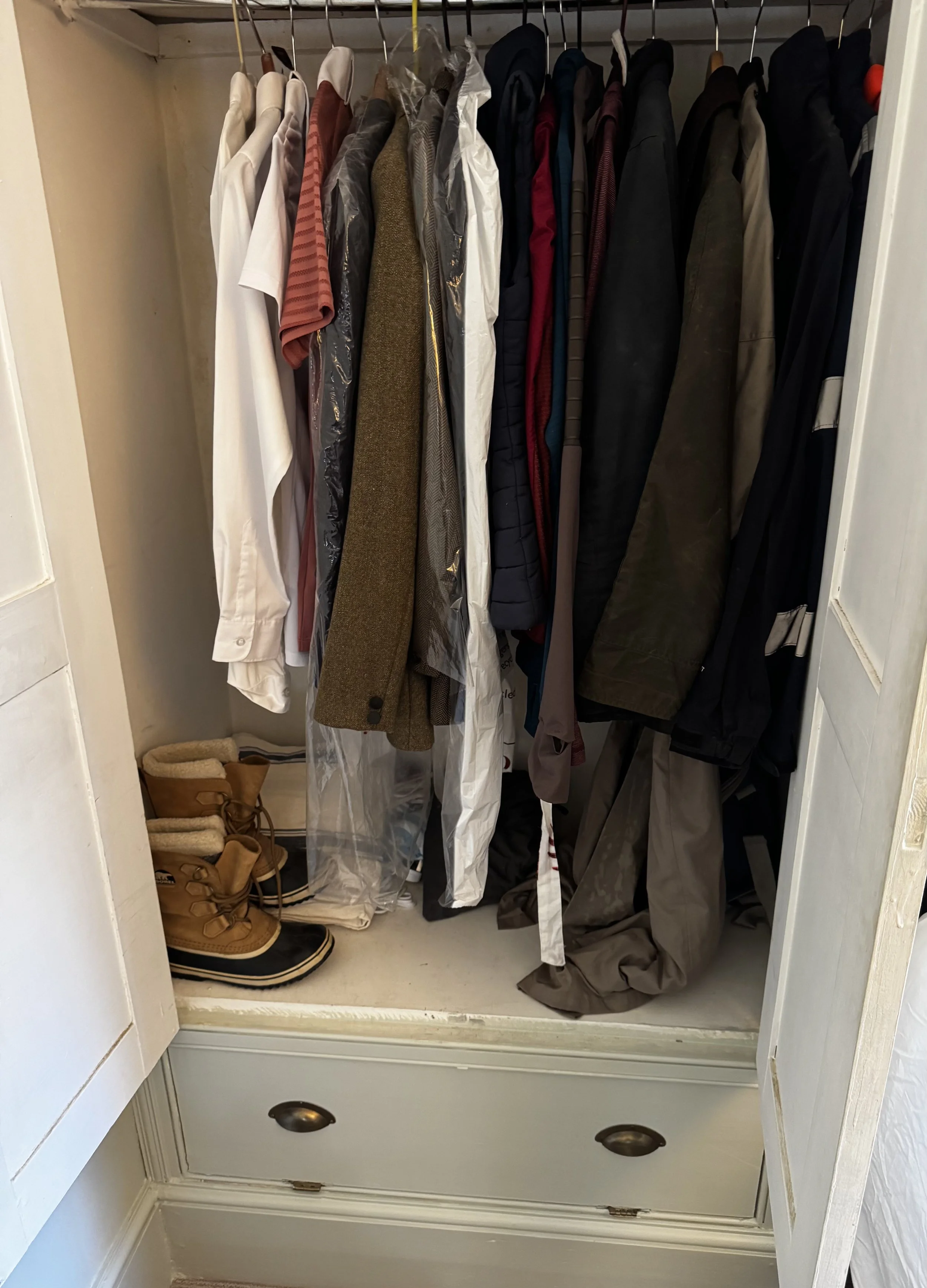 Inside a closet with hanging clothes including shirts, jackets, and coats. Two pairs of tan winter boots are on the closet floor.