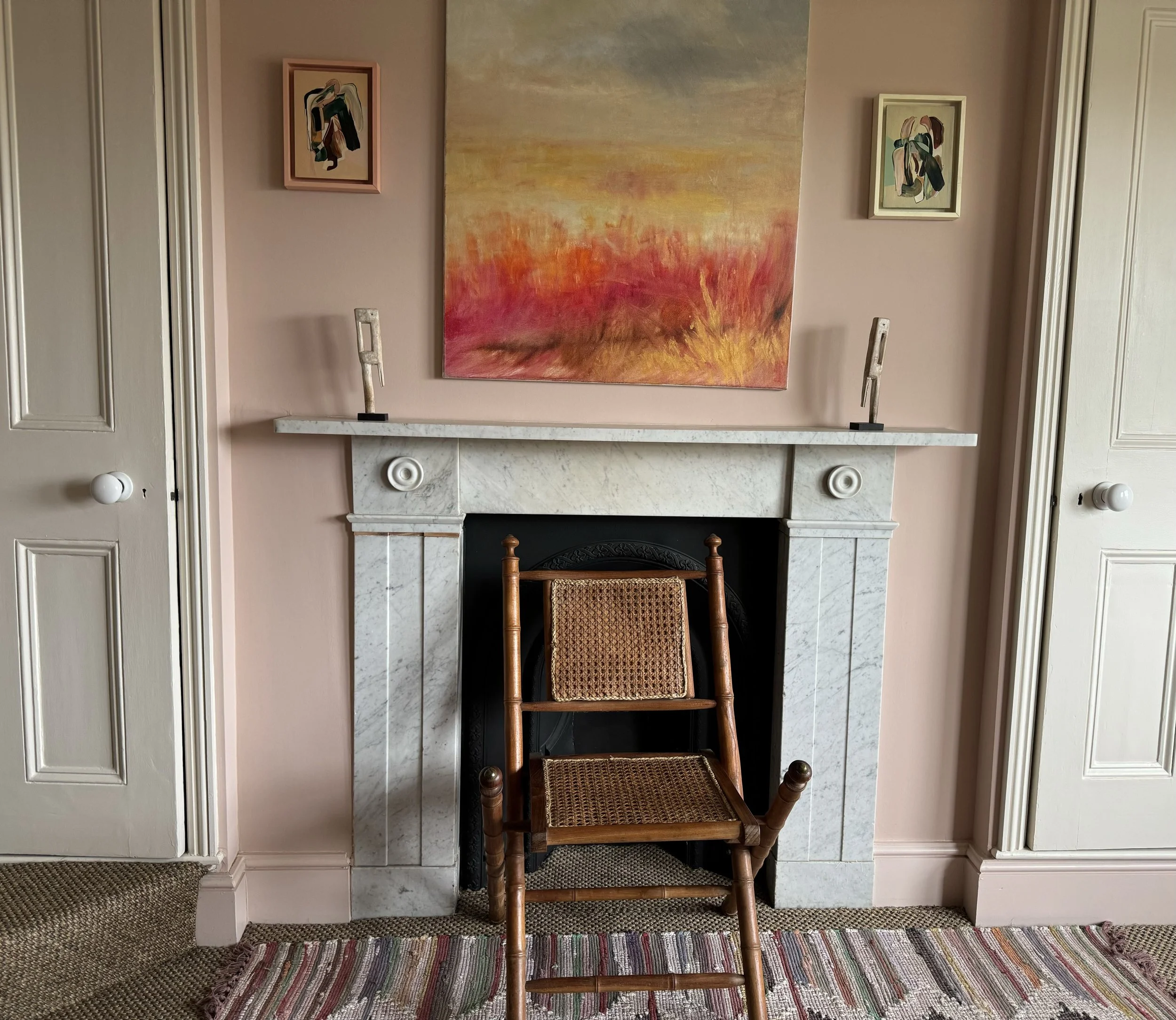 A cozy living room corner with a white marble fireplace, framed abstract paintings above and on sides, a wooden chair with woven seat in front, and a colorful striped rug on the floor.
