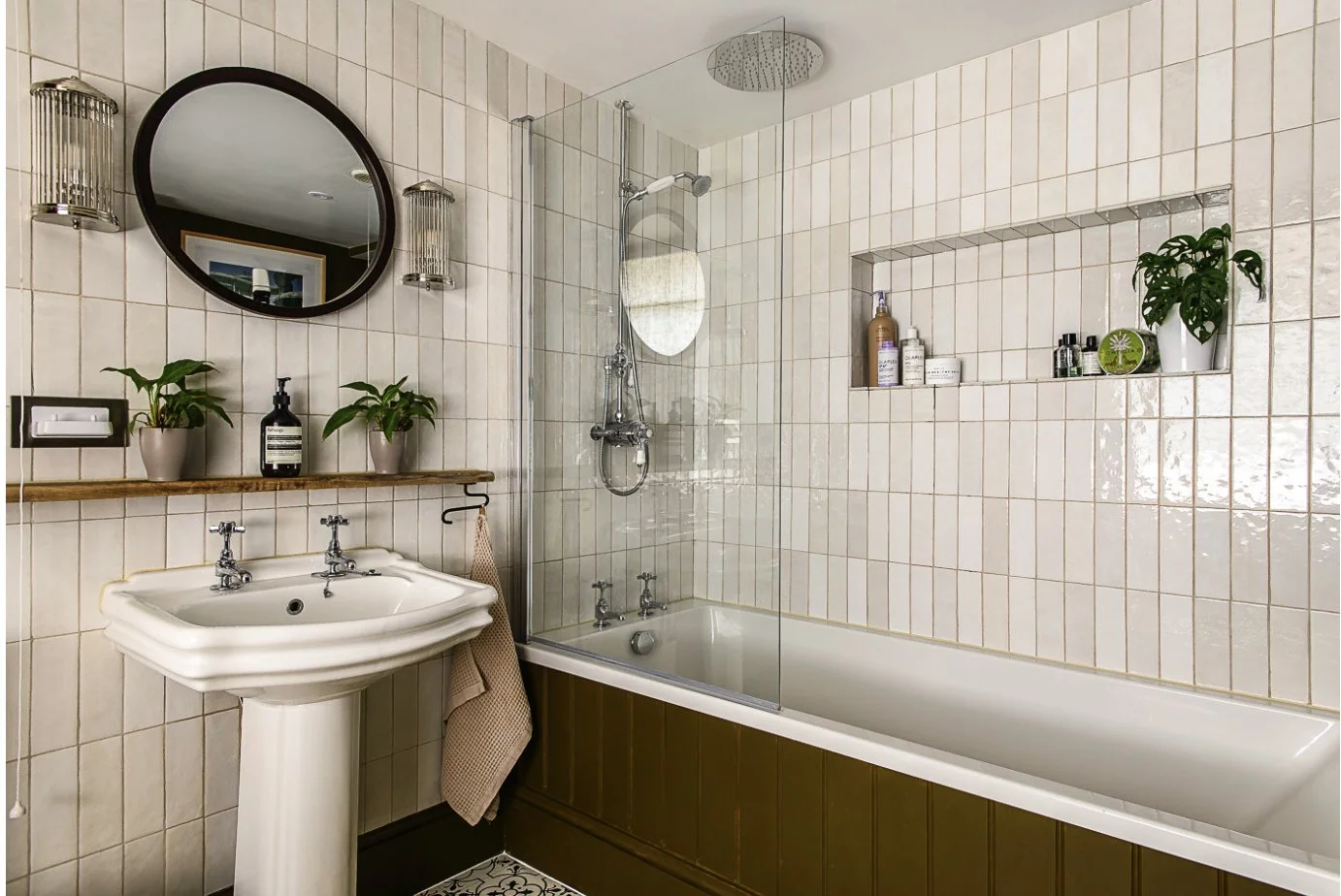 A bathroom with beige tiled walls, a white bathtub with a brown panel, a white pedestal sink, a round mirror, and potted plants on shelves.