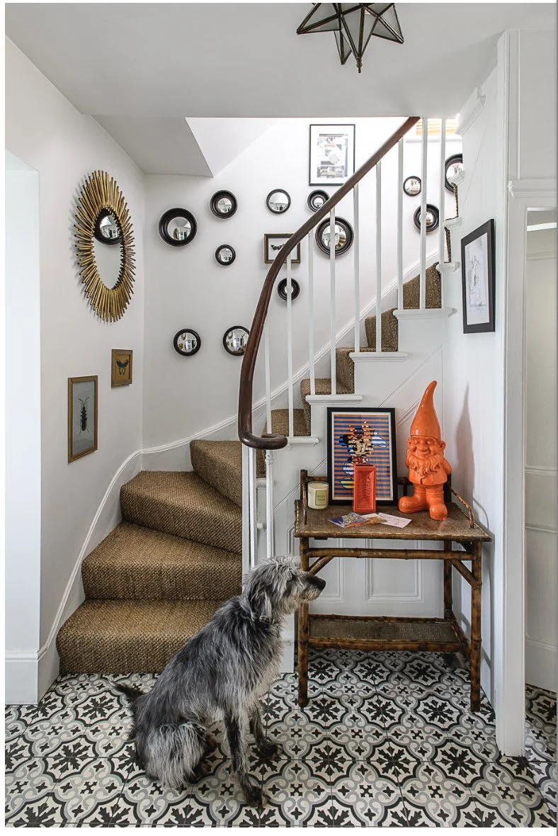 A small indoor entryway with a staircase, decorative wall mirrors, framed pictures, a table with a gnome statue, candles, and artwork, and a dog sitting on patterned tile floor.