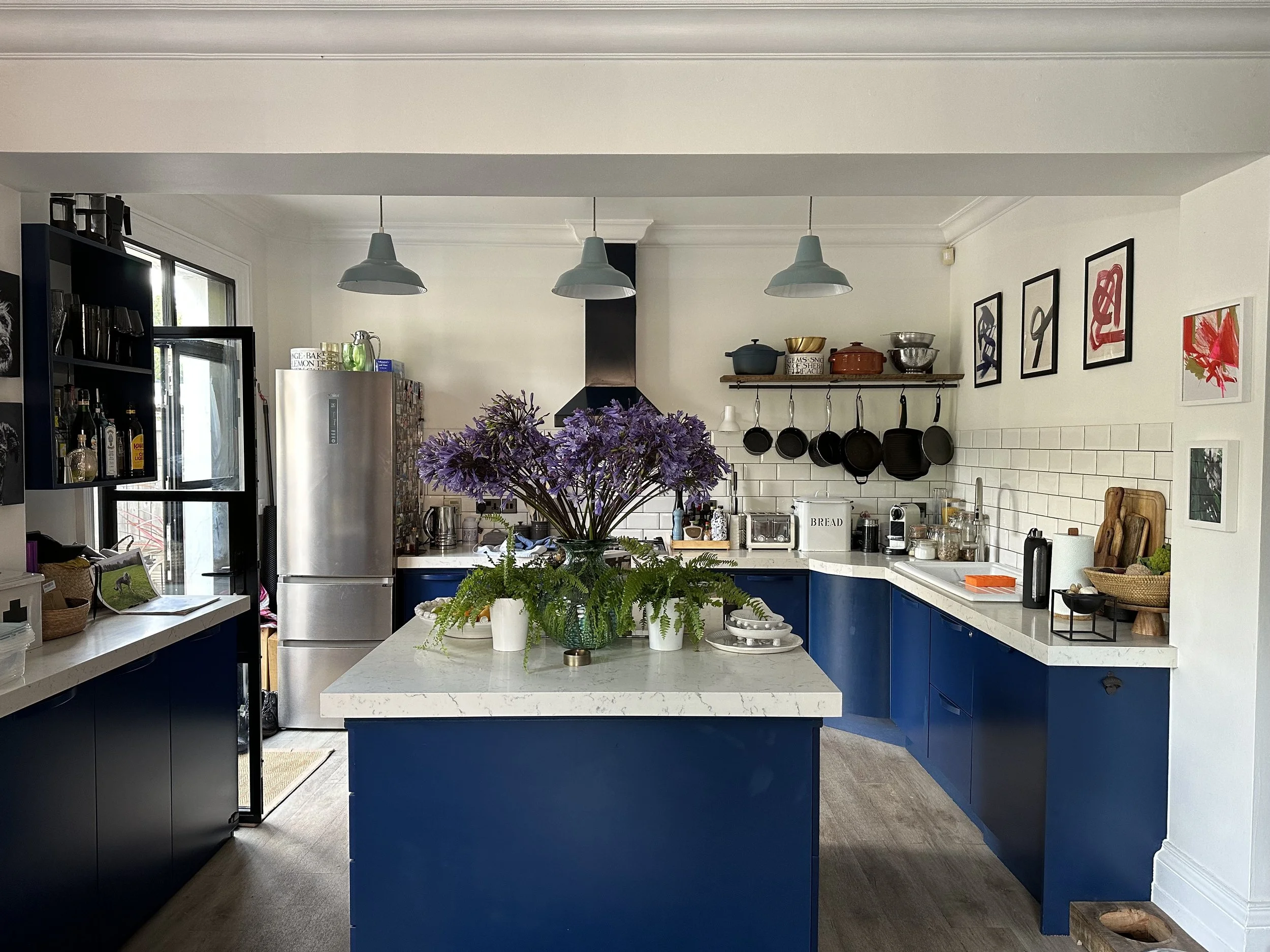 Modern kitchen with white countertops, dark blue cabinets, a center island with flower arrangement, open shelving with pots, framed artwork, and appliances including a stainless steel fridge and microwave.