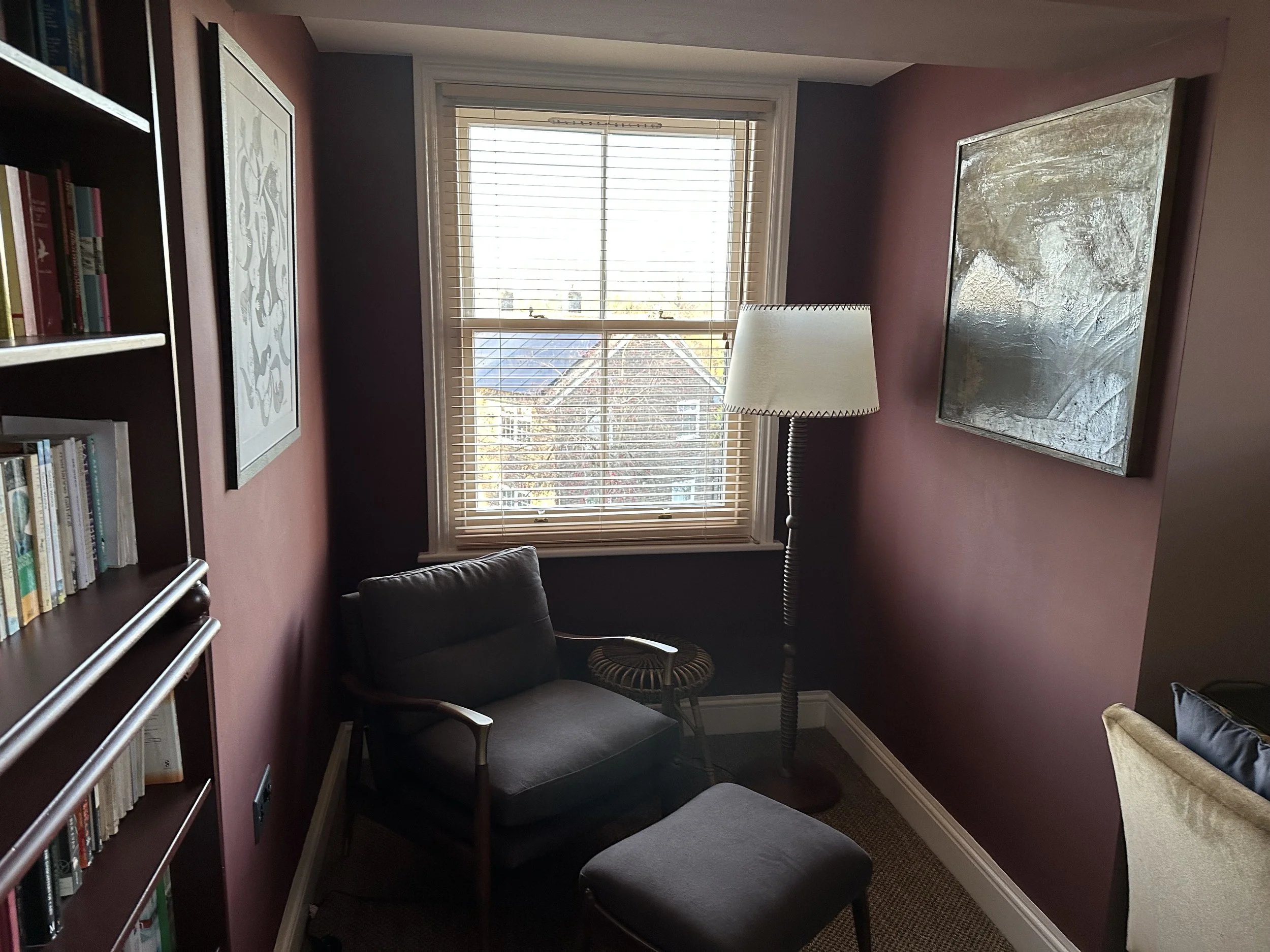 A cozy corner of a room with a window, a dark armchair with a matching footstool, a tall floor lamp, bookshelf, and framed artwork on purple walls.