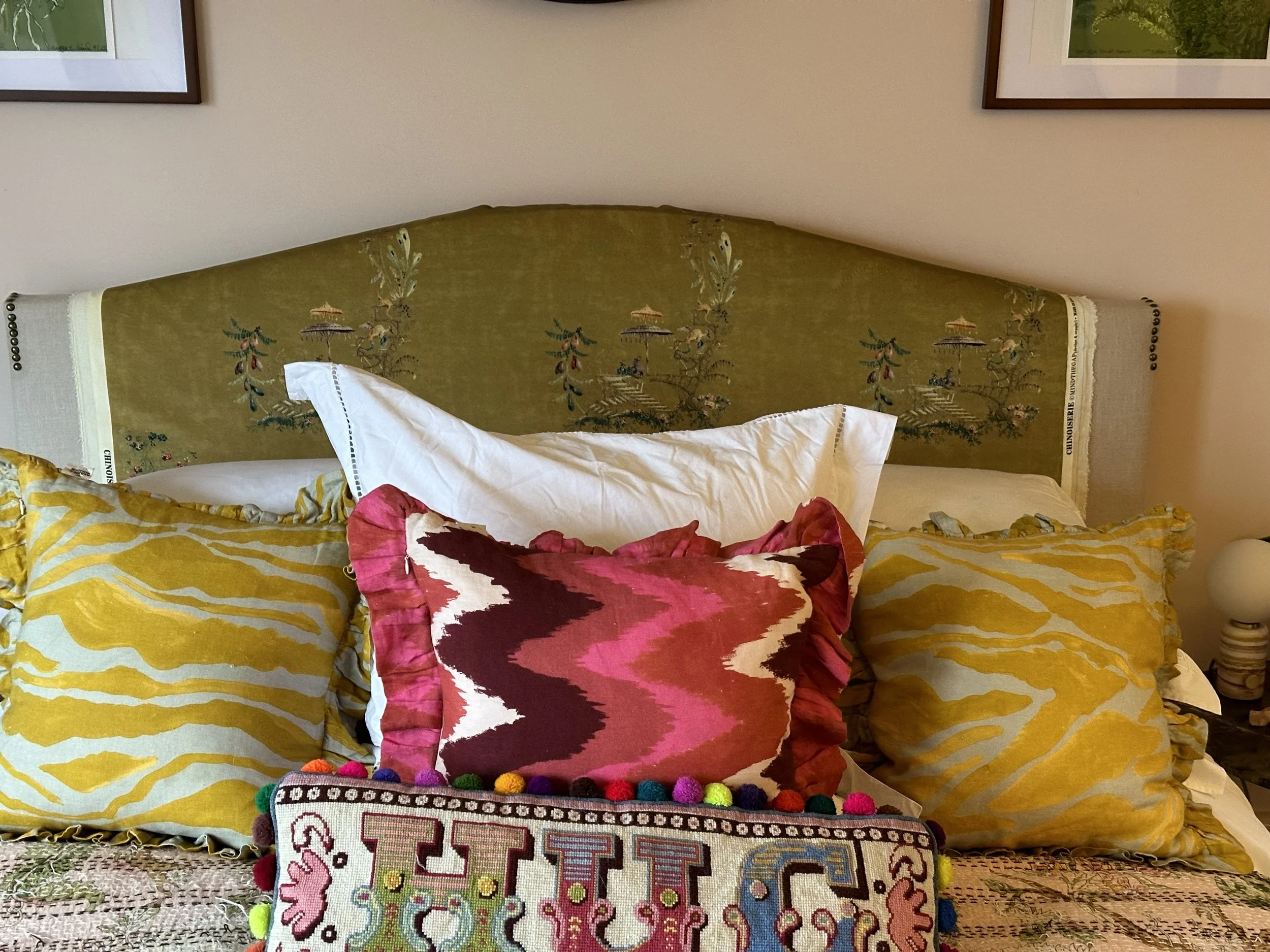 A bed with a green vintage-style headboard featuring embroidered scenes, yellow patterned pillows, a decorative pink, red, and white zigzag pillow, and a colorful embroidered textile with pompoms in front.