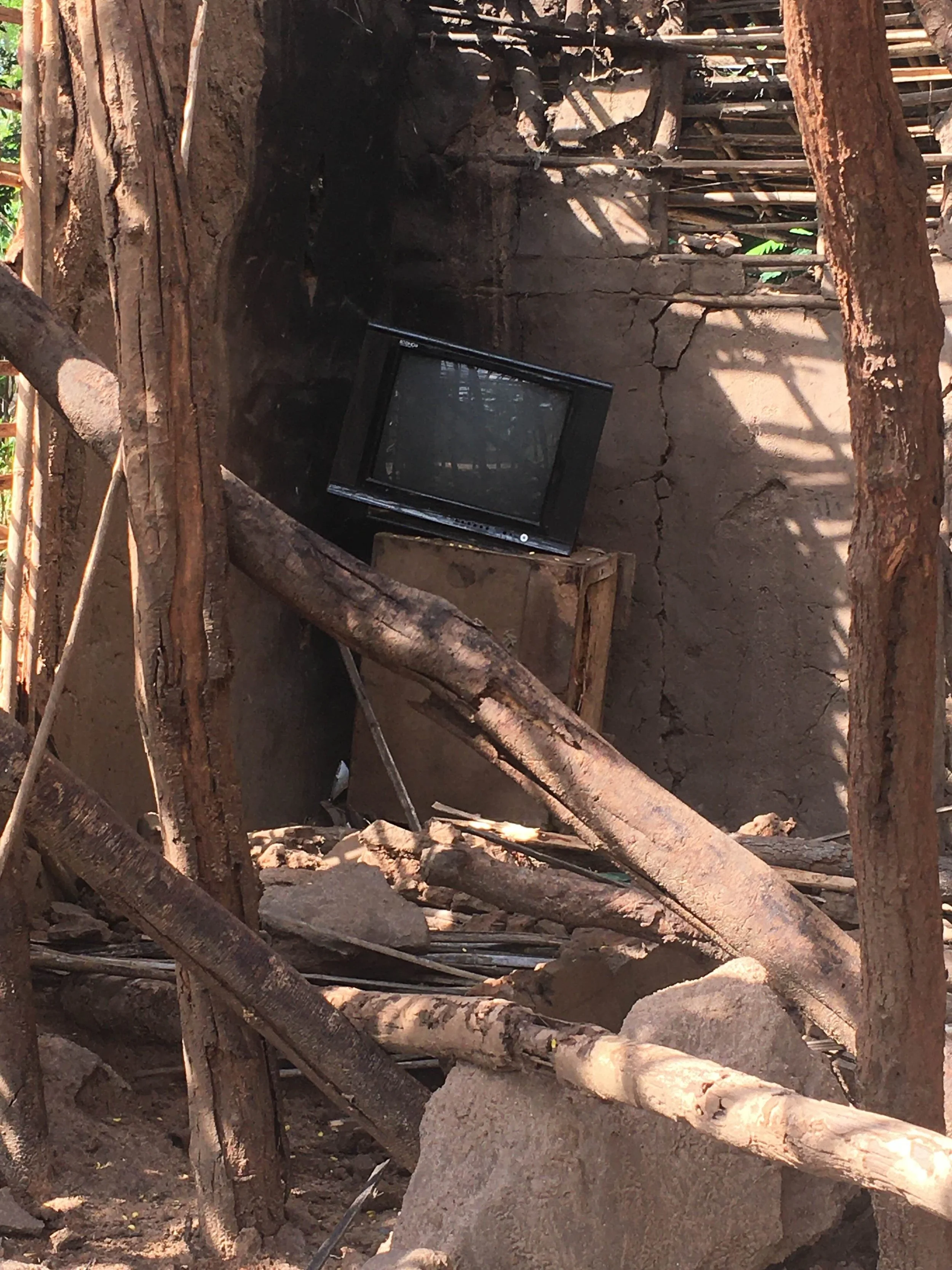 An old, broken television resting on a makeshift wooden platform amidst debris and tree branches, with a background of a cracked clay wall and shadows.