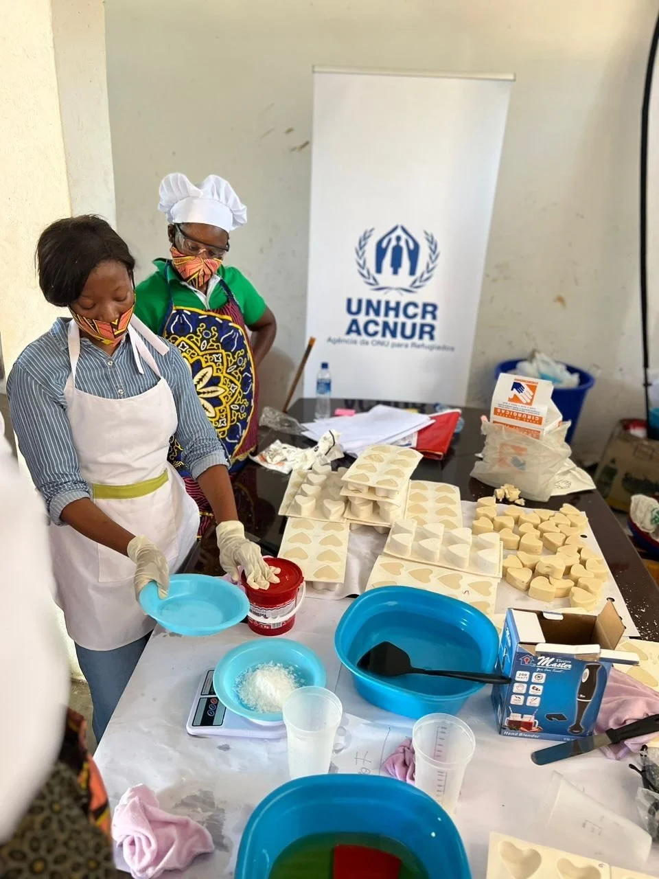 Two women in colorful clothing and masks, wearing aprons and gloves, preparing soap in a room with a UNHCR banner in the background. The table is filled with soap molds, ingredients, and tools.