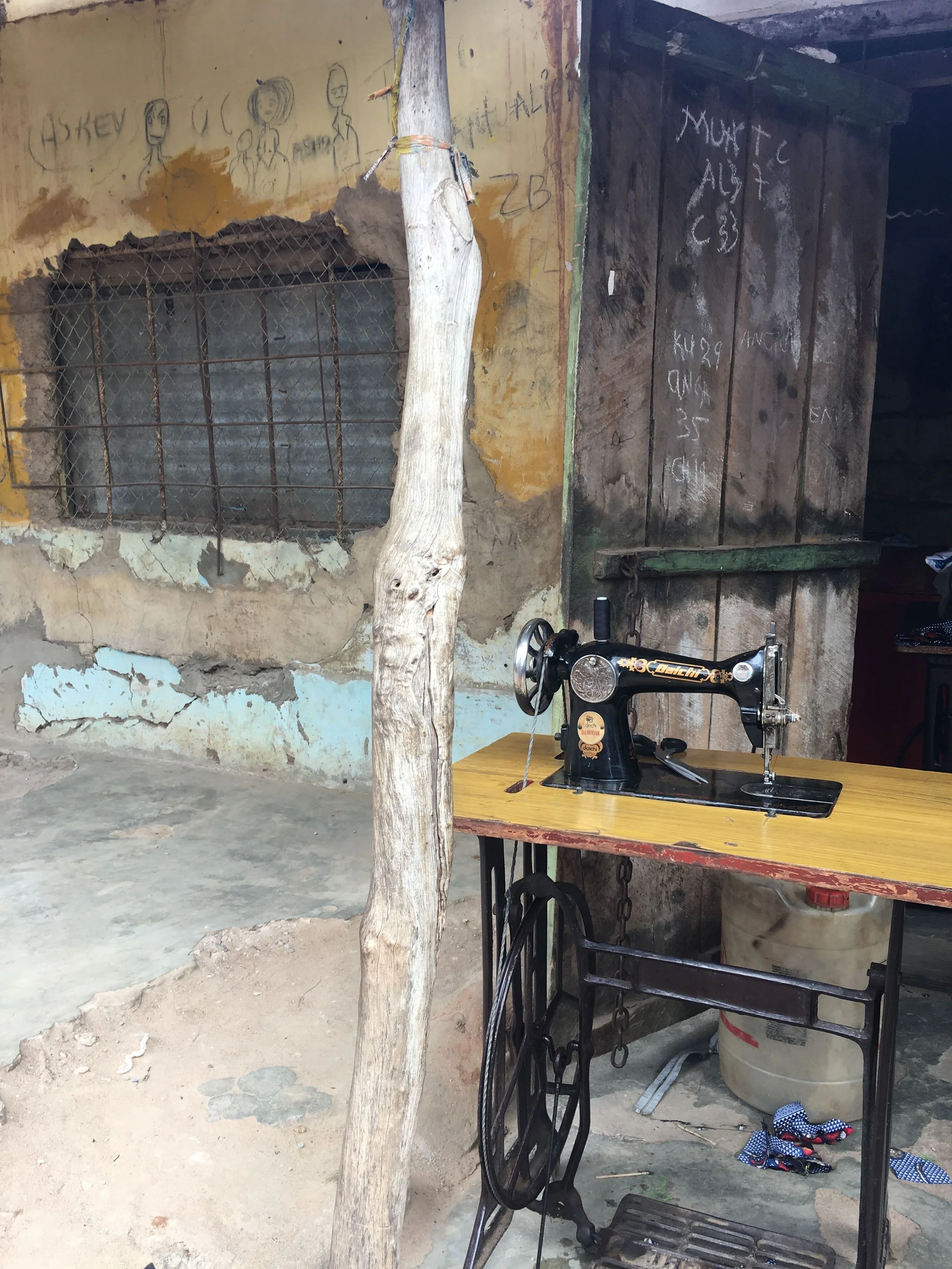 An old sewing machine on a yellow wooden table in a rustic, worn-out room with weathered walls, a boarded-up window with metal bars, and a rough chain and wooden support. Scattered items include a plastic container and some cloth on the floor.