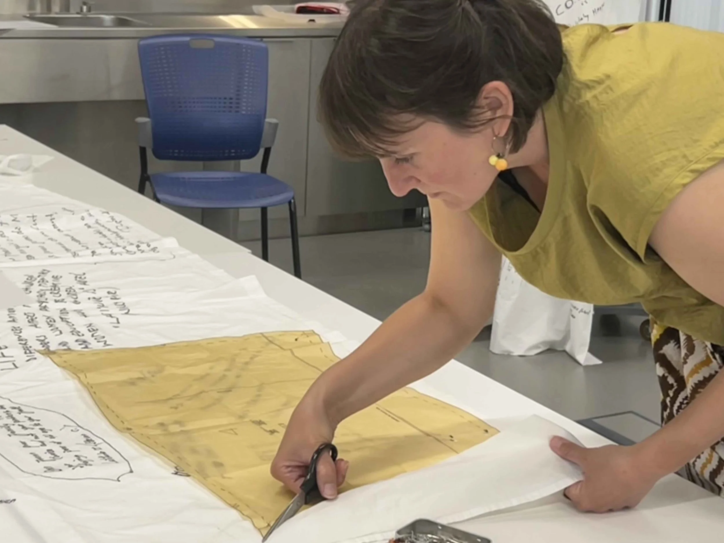Woman cutting a large handwritten paper with scissors at a table in an indoor setting.