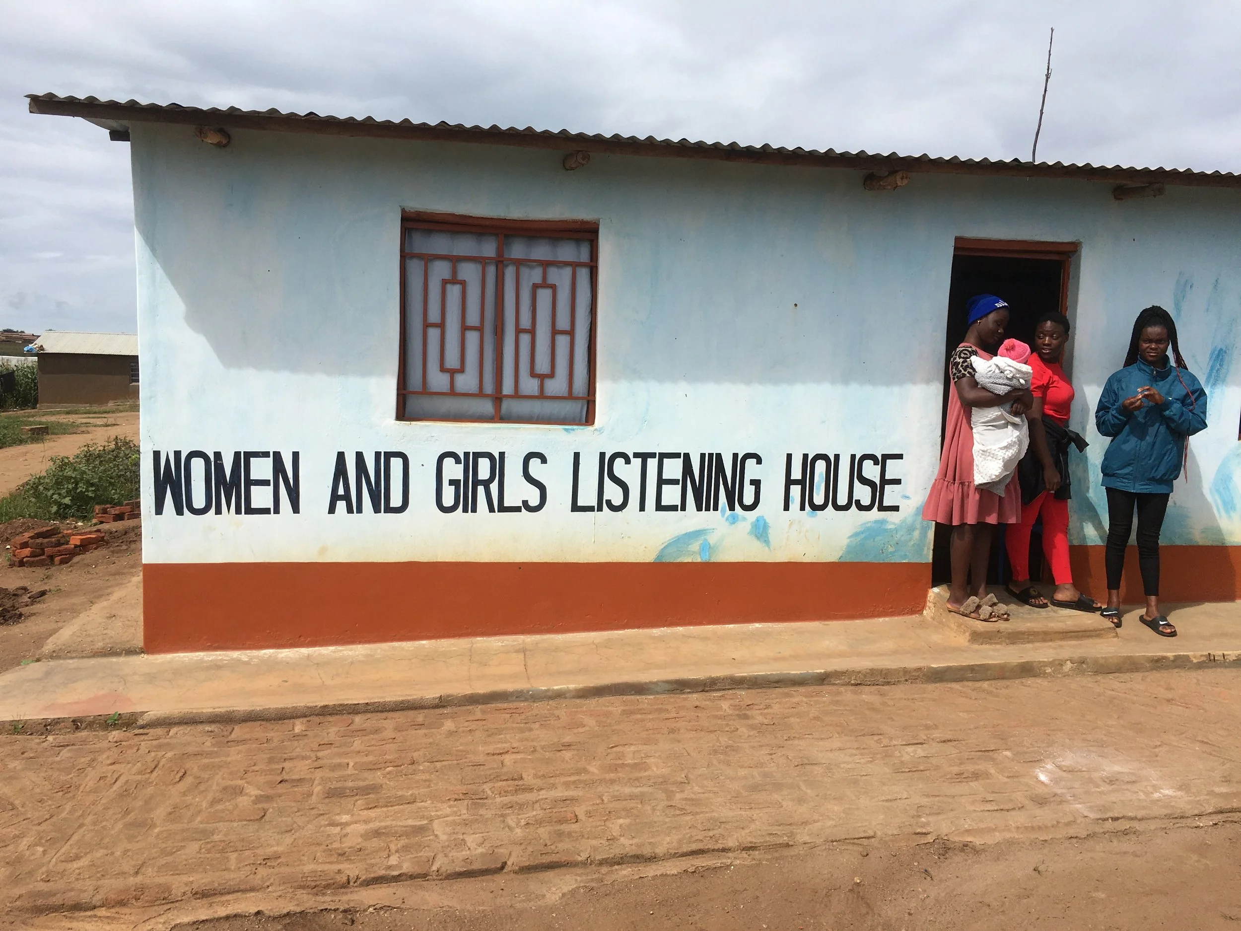 A building with a sign that reads 'Women and Girls Listening House' and three women standing outside, one holding a baby.