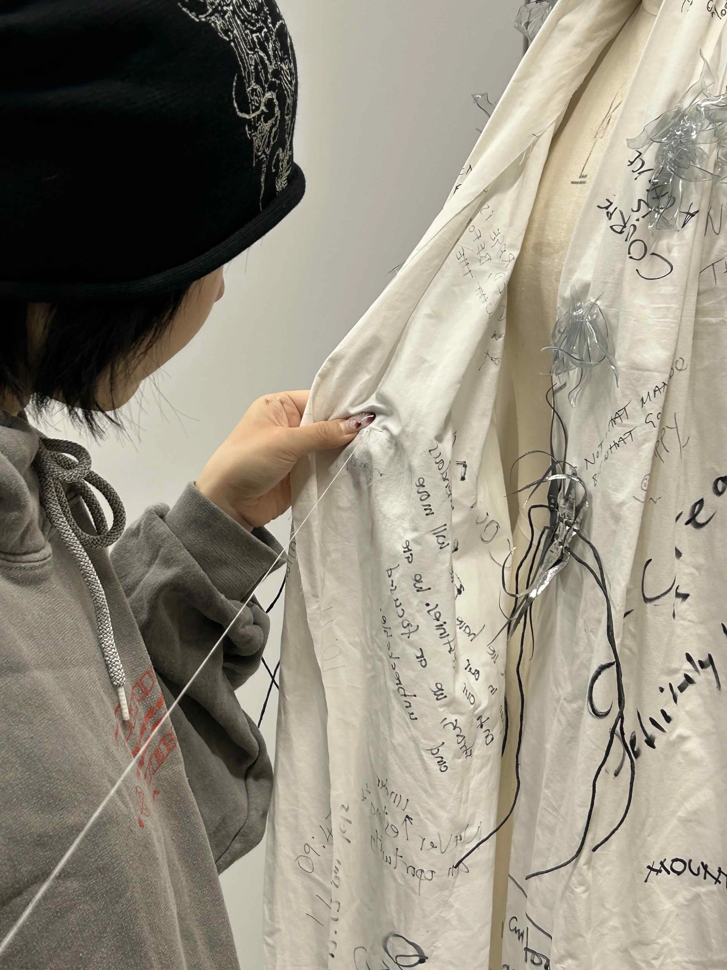 Person wearing a black beanie and gray hoodie examining a fabric with handwritten notes and drawings. The fabric is white or cream-colored with black ink writing and some decorative silver elements.