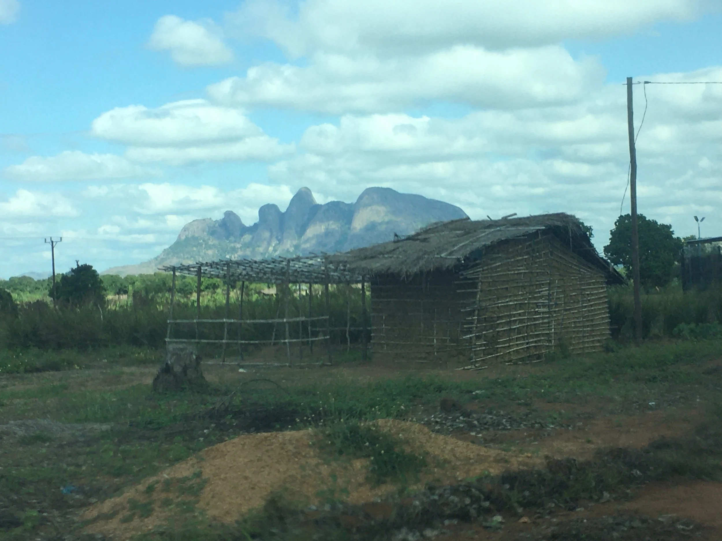 A rural landscape featuring a thatched hut made of wooden sticks, a dirt road, power poles, and a mountain range in the background under a partly cloudy sky.