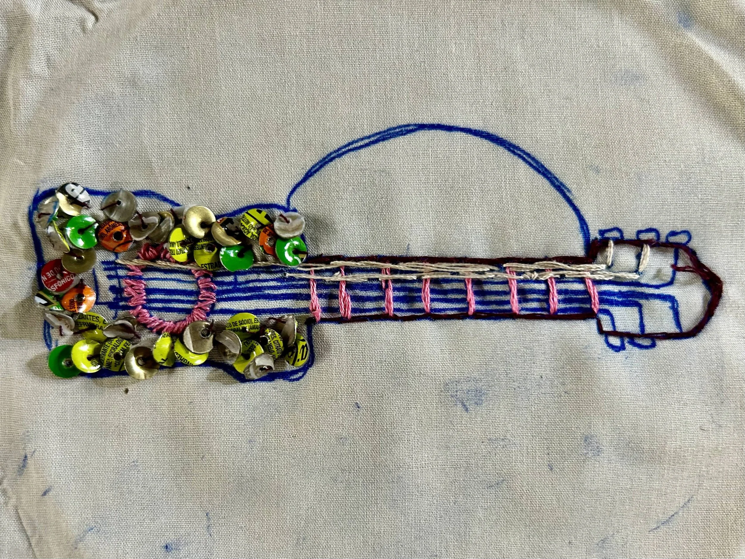 An embroidered design of a guitar on fabric, outlined with blue thread. The guitar's body is decorated with yellow and black debris, possibly bottle caps, and the neck is stitched with pink thread. The sketch of the guitar is outlined with blue threa
