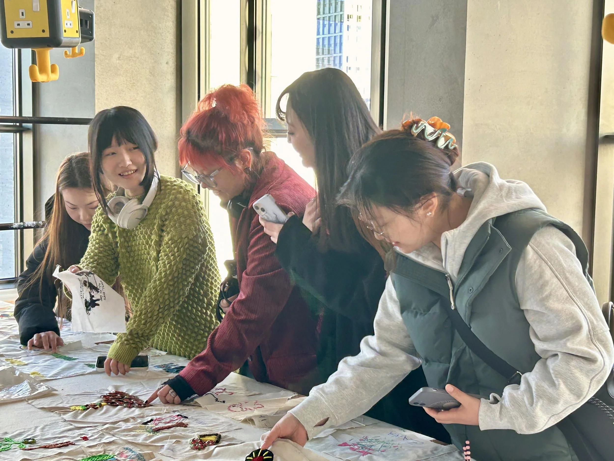 A group of five women gathered around a table, looking at handcrafted items, in a brightly lit room with large windows.