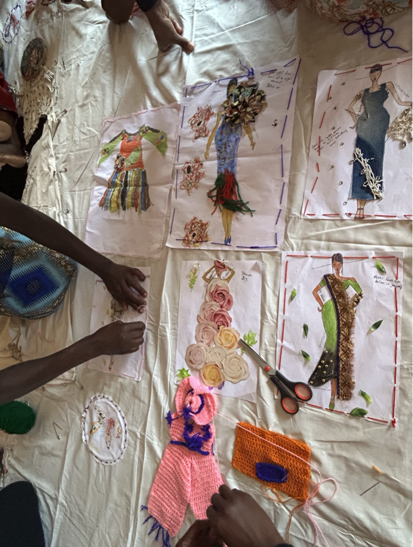 People working on fashion illustrations, cutting and arranging fabric and paper designs for clothing, with scissors, thread, and decorative elements on a table.